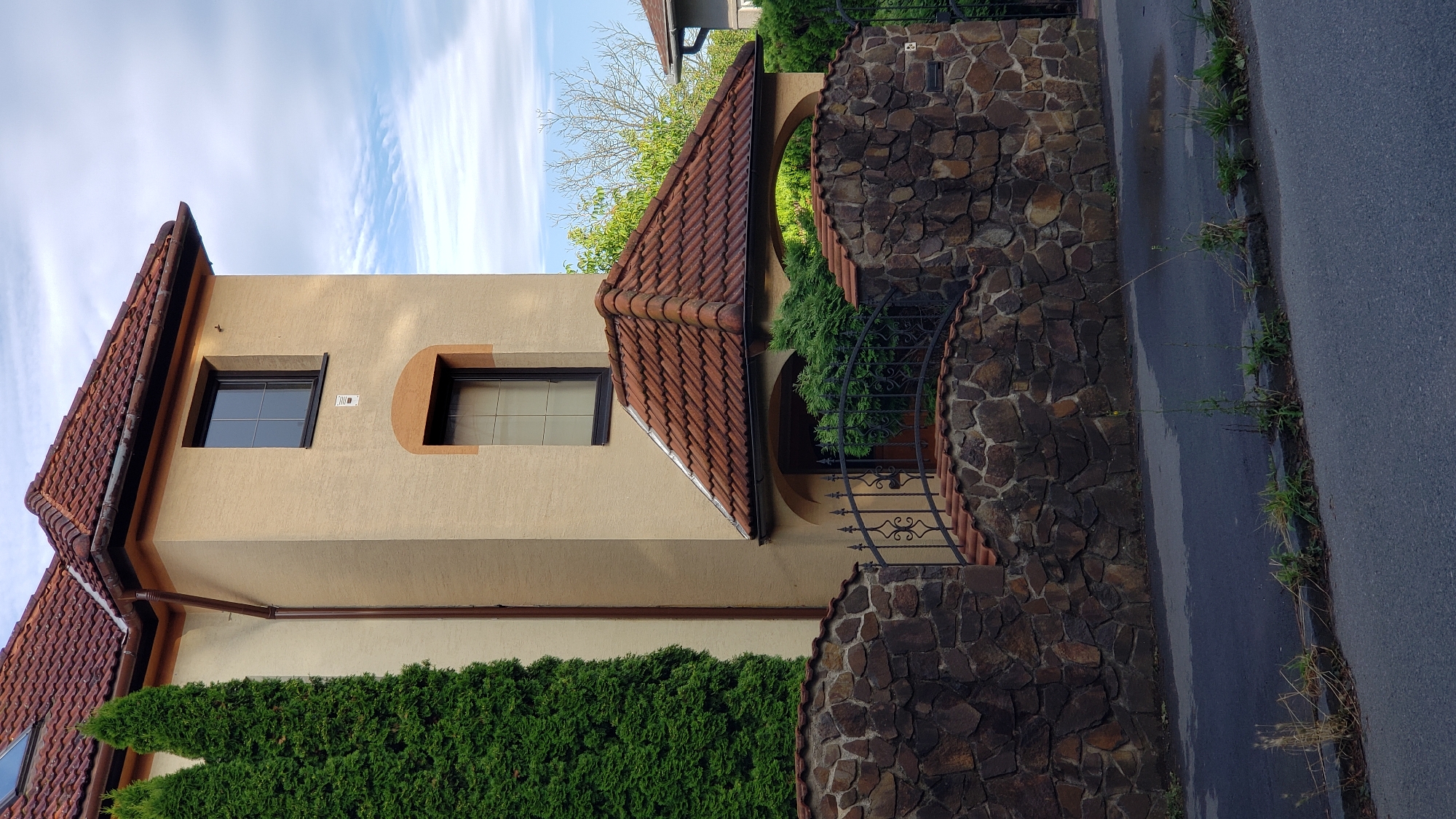 This image captures a section of a residential building in Prešov, Slovakia. The building features a light beige stucco exterior with a red tiled roof. A prominent architectural element is a tall, narrow tower-like section of the building, accented with a curved, lighter beige trim around an upper window. There are two visible windows on this tower section, both with dark frames and mullions. To the left of the tower, a tall, dense evergreen tree stands adjacent to the building's wall.
In the foreground, a decorative stone wall constructed from irregular, dark-colored stones forms a barrier. A black wrought-iron fence with an ornate, curved top runs along the top of the stone wall, marking an entrance or boundary. A small, pitched red-tiled roof extends from the building over an entryway, supported by arched openings. Green shrubbery is visible behind the fence and beneath the entryway roof.
The street in front of the property is paved with asphalt. A curb separates the street from a narrow strip of grass and weeds. The sky above is partly cloudy with streaks of white clouds against a blue background, suggesting daylight conditions. The overall scene depicts a well-maintained residential property.