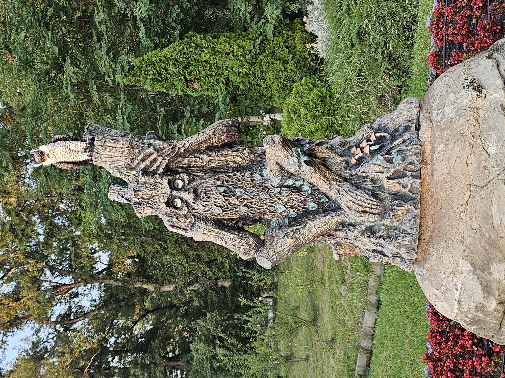 The image depicts a statue resembling a wise old tree spirit with a long, tangled beard and wide, staring eyes. The statue is made to look like weathered wood and is adorned with small mushrooms. Perched atop the statue is a realistic-looking owl. The statue stands on a large, rounded stone base, surrounded by vibrant red flowers and lush green grass. In the background, a dense forest with various trees and a tall evergreen tree creates a natural, park-like setting. The scene is lit by natural daylight, suggesting it was taken outdoors during the day. This statue is located in Cherkessk, Russia.