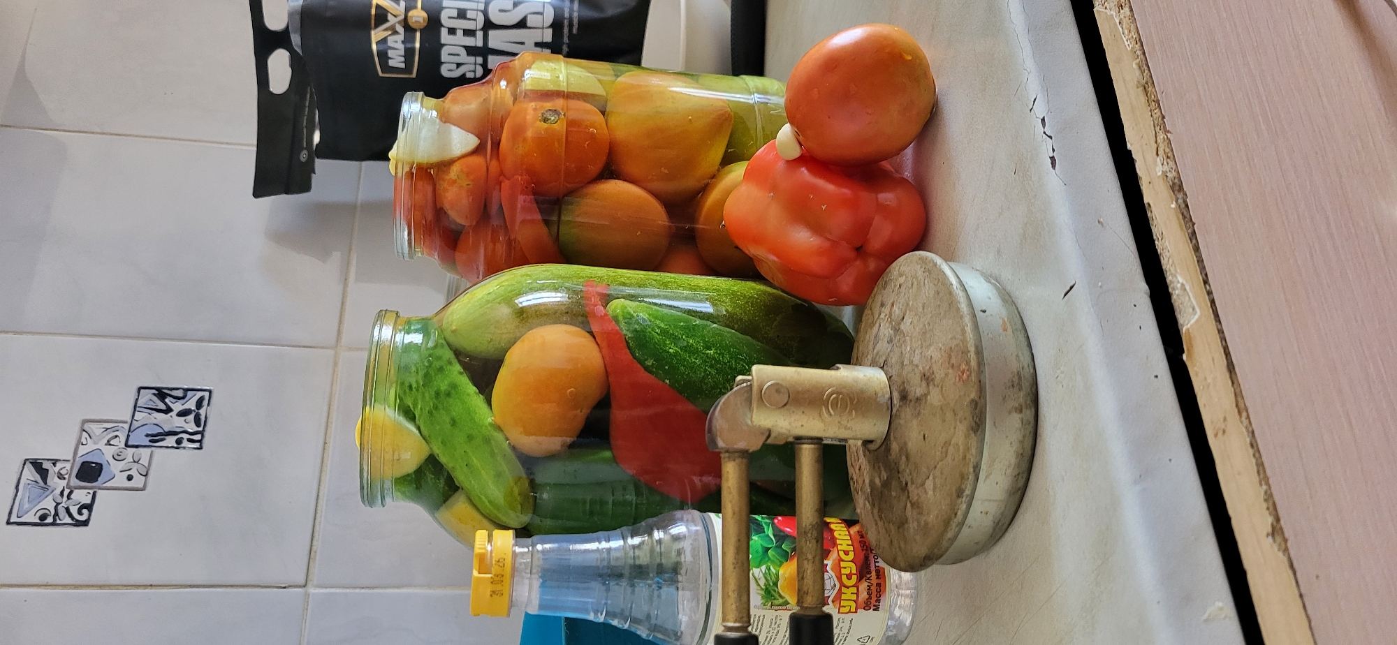 A close-up, eye-level shot captures a countertop scene in what appears to be a kitchen in Oskemen, Kazakhstan. The foreground is dominated by a tarnished, circular metal object with a brass fixture attached, reminiscent of a small stove or burner. To the left of this object, a clear glass bottle with a yellow cap and a label featuring images of fruits and text, likely containing vinegar, stands upright. Behind these items, two large glass jars are prominently featured. The left jar is filled with pickled cucumbers, interspersed with slices of lemon and what appear to be small orange fruits and red peppers. The right jar contains pickled tomatoes and slices of lemon. To the far right, a red bell pepper and a tomato rest on the countertop. In the background, a white tiled wall serves as the backdrop, with three square decorative tiles arranged vertically on the upper left section. A black bag with the word "MAX" and "SPECIAL" visible is partially visible on the right side of the frame. The overall impression is one of domestic food preservation.