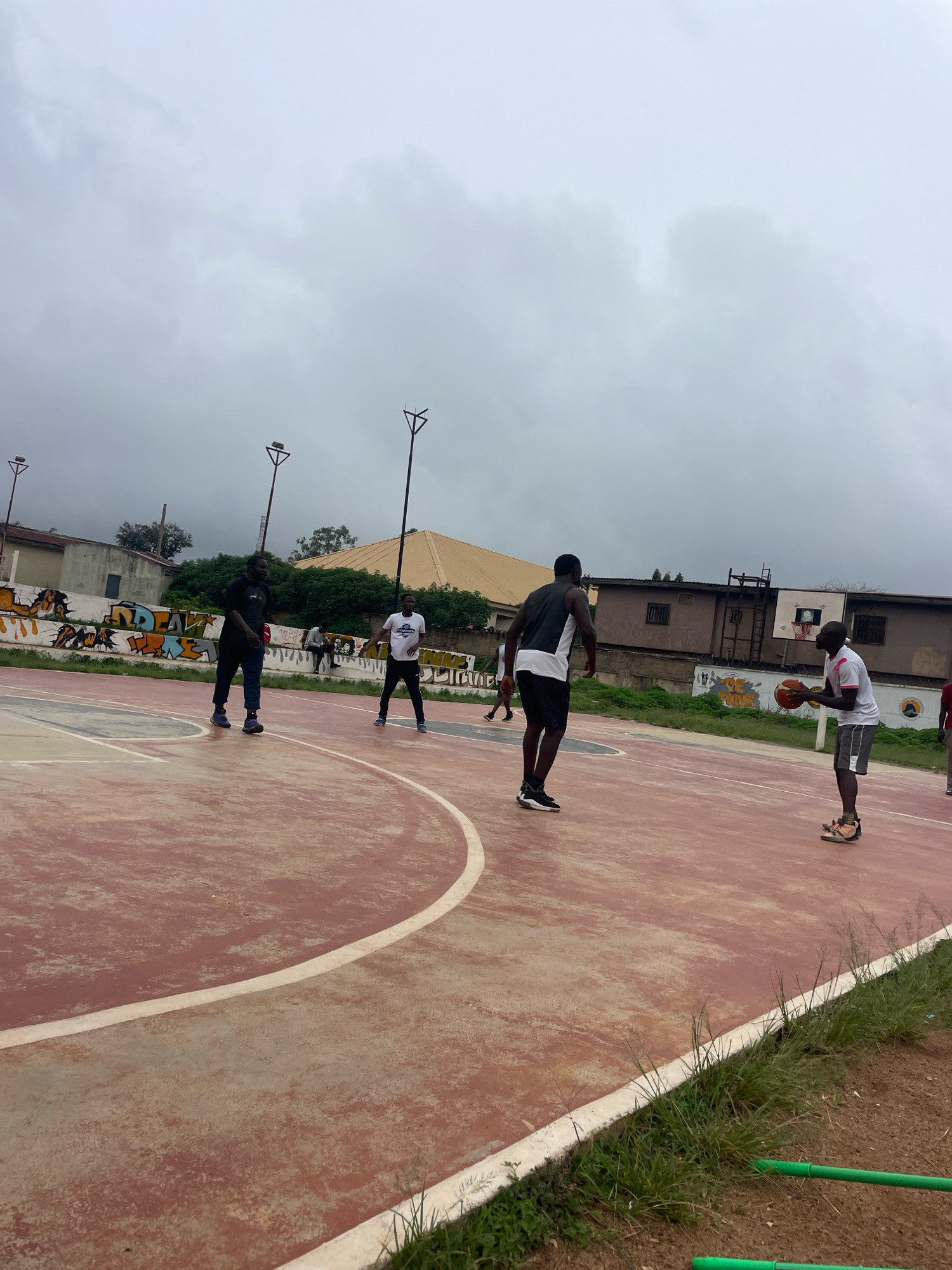 The image depicts an outdoor basketball court on a cloudy day in Gura Top, Nigeria. Several individuals are engaged in a game of basketball. In the foreground, a person in a white shirt and grey shorts holds a basketball, appearing ready to pass or shoot. Opposite them, another person in a black and white sleeveless shirt and black shorts stands defensively. Further back, two other individuals are visible on the court. One, wearing a black t-shirt and dark pants, stands with hands on hips. Another, in a white t-shirt and dark pants, is also on the court. The court itself is reddish-brown with white markings. In the background, a wall adorned with graffiti runs along the perimeter of the court, beyond which are buildings and trees. Two tall light poles are also present, suggesting the court may be used in the evening. The overall environment appears somewhat worn, with grass and dirt surrounding the court.
