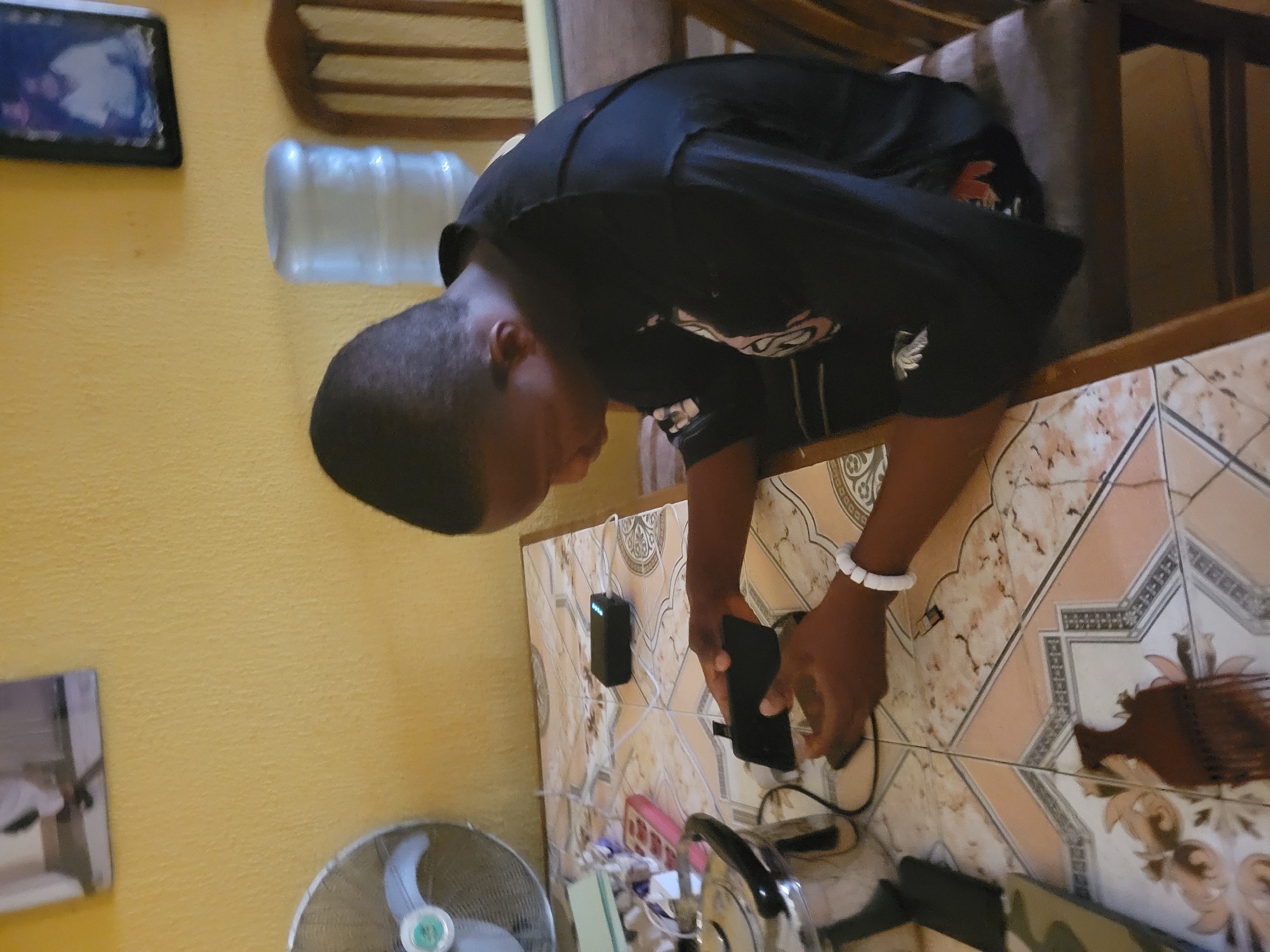 The image depicts a young Black male seated at a table covered with a tiled surface, in Onitsha, Nigeria. He is wearing a black t-shirt and a white bracelet on his left wrist. His attention is focused on a black smartphone he is holding in his hands. Nearby on the table are a black power bank with indicator lights illuminated, and a power strip with multiple outlets, some of which are plugged in. A metal pitcher with a black handle is also visible on the left side of the table. The background shows a yellow wall adorned with two framed pictures. To the right of the male, a portion of a wooden chair and a large water dispenser are visible. A floor fan is positioned on the far left of the table. The overall setting suggests an indoor domestic environment.