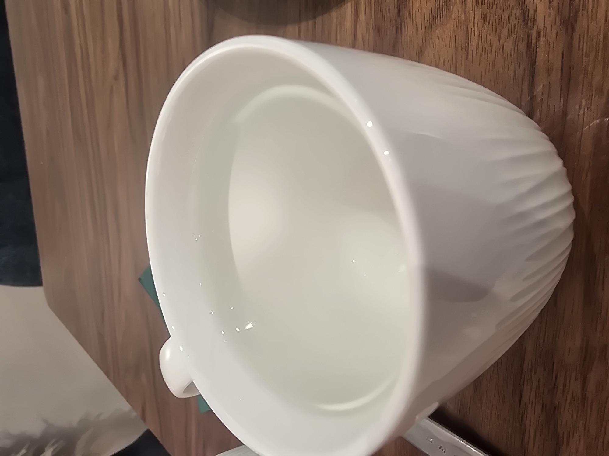 A white, ribbed ceramic bowl filled with clear liquid sits on a wooden surface. The bowl is tilted, with its opening facing the viewer. The liquid reflects light in small, bright spots. The wooden surface appears to be a tabletop, with visible grain patterns. A dark green object is partially visible beneath the bowl, and to the left of the bowl, a lighter colored surface with a blurry pattern is seen. The lighting suggests an indoor setting, possibly during the day. No people are visible in the image. There is no visible text.