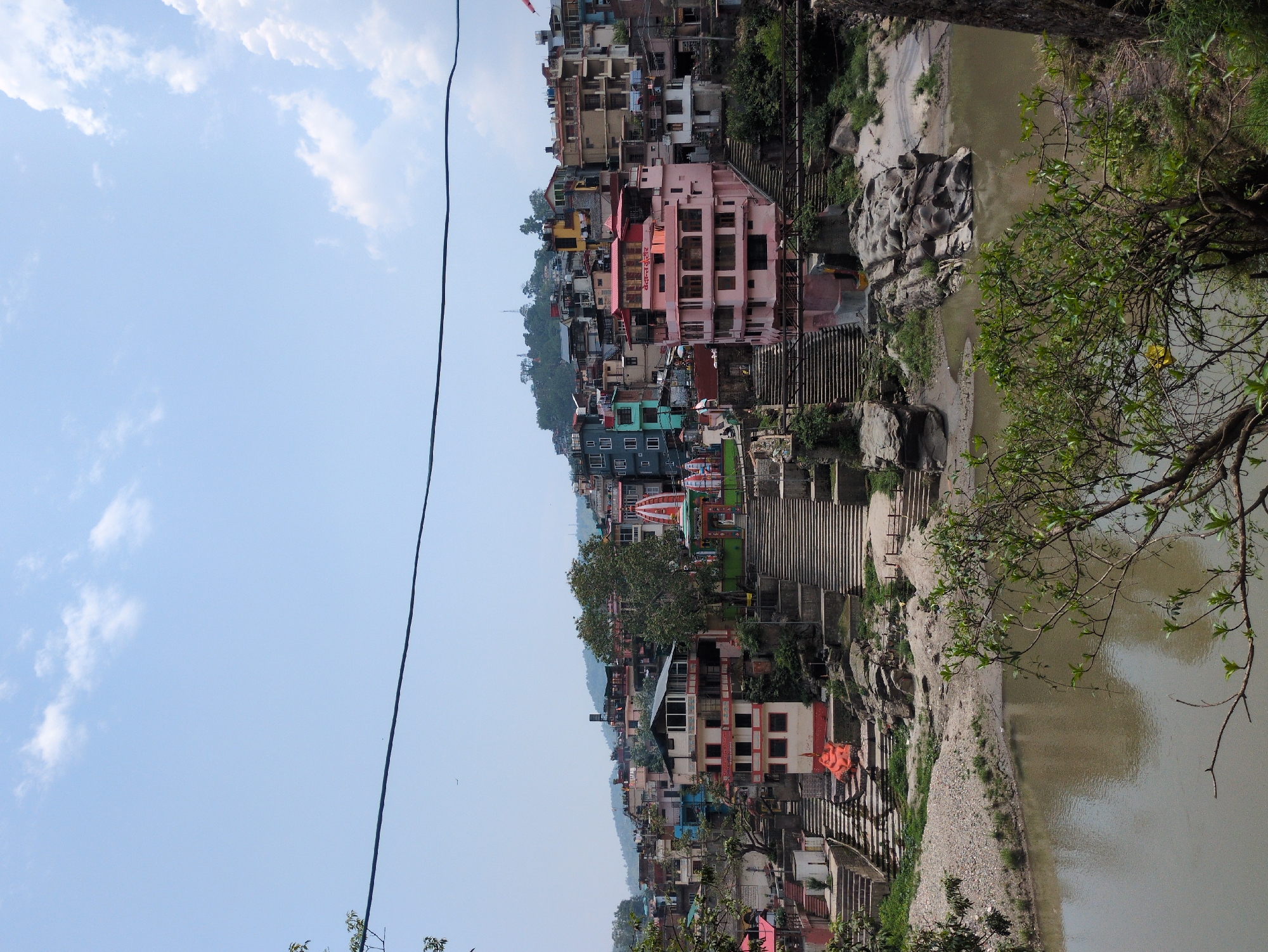 The image depicts a cityscape nestled beside a river, likely in the Indian city of Mandi, known for its temples and hilly terrain. The central focus is a densely packed collection of buildings constructed in a stepped, terraced fashion up a hillside. The architecture is varied, with structures featuring colorful facades—pinks, blues, whites, and yellows—and pitched roofs. Many buildings appear to be residential or commercial, with multiple stories and windows.
In the foreground, a river flows, its surface relatively calm and reflecting the muted tones of the sky and surrounding structures. The riverbanks are a mix of rocky outcrops, gravelly areas, and some sparse vegetation. Several sets of stone stairs descend from the town towards the river, suggesting pathways and access points.
Prominent in the mid-ground is a large, pink building with a distinctive stepped facade and a red-tiled roof. To its left, a vibrant orange statue of a deity is visible, situated near the base of a wide set of stone stairs. Further to the left, a red and white structure with an ornate, pagoda-like roof (likely a temple or shrine) stands, with smaller buildings surrounding it. In the upper part of the image, the sky is a light blue, partially obscured by scattered white clouds. The overall impression is one of a vibrant, densely populated area situated in a natural landscape, likely a religious or historical center given the presence of stairs and structures resembling temples. A single power line traverses the upper portion of the frame, indicating modern infrastructure within the scene.