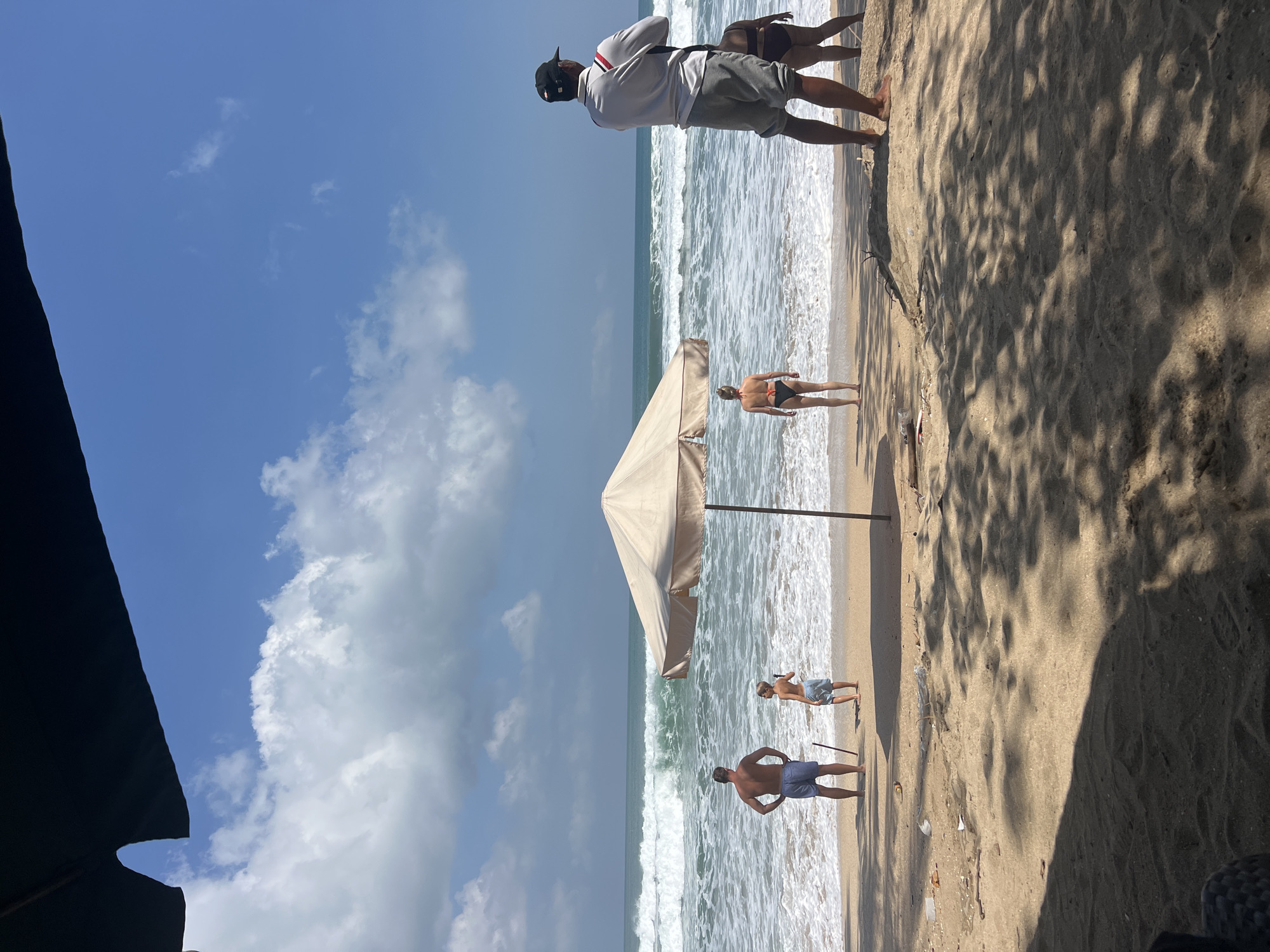 The image depicts a sunny day at Kuta Beach, Indonesia. The scene is viewed from under a dark canopy, likely an umbrella or awning, casting dappled shadows on the sandy foreground. In the middle ground, a large, beige beach umbrella stands open. Several people are visible: a man and a boy stand near the water's edge, facing the ocean; a woman in a bikini is also facing the sea. To the right, a man wearing a cap, grey shirt, and grey shorts stands facing away from the camera, looking out at the ocean. Another person, partially obscured, is standing next to him. The ocean is active with moderately sized waves crashing on the shore. The sky is blue with scattered white clouds. Scattered debris, including plastic and sticks, is visible on the sand near the water's edge.