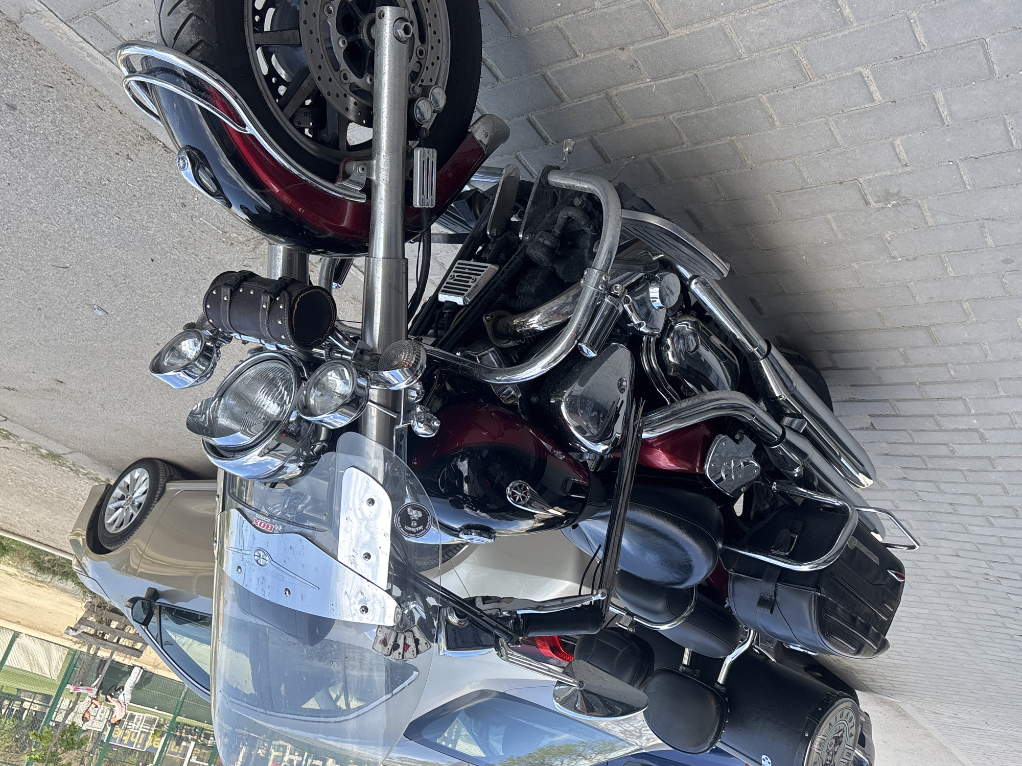 A large, classic-style cruiser motorcycle, featuring a striking red and black paint scheme and extensive chrome detailing, is prominently parked outdoors in Plovdiv, Bulgaria. The image, captured during clear daytime under bright sunlight, shows the motorcycle resting on a paved surface composed of grey brick pavers to its right and a lighter concrete or asphalt area to its left.
The front of the motorcycle boasts a large central headlight, flanked by two smaller auxiliary lights, all encased in shining chrome. A black, cylindrical tool bag adorned with studs is mounted below the headlights. A clear windshield with chrome accents protects the handlebars; a red label with the text "BOX" is visible on its inner side, alongside a "Jack Daniel's" sticker. The front wheel, equipped with disc brakes, and the red and black front fender complete the motorcycle's leading features.
Towards the mid-section, the glossy red and black fuel tank catches the light. The engine block features chrome covers, including one embossed with "YAMAHA," leading to polished chrome exhaust pipes. Black leather seats for both rider and passenger are present.
The rear of the motorcycle is fitted with black leather saddlebags on either side, supported by chrome frames. At the very back, a large, round black storage container, possibly a trunk, displays a decorative chrome plate with a compass-like design and partially visible numbers, including "12" and "96." Chrome crash bars are integrated around the motorcycle's engine and rear sections.
In the background, a light-colored sedan is partially visible behind the motorcycle. To the left, a green metal fence borders a grassy area, suggesting the motorcycle is parked near a park or a green public space. No individuals are present in the scene.