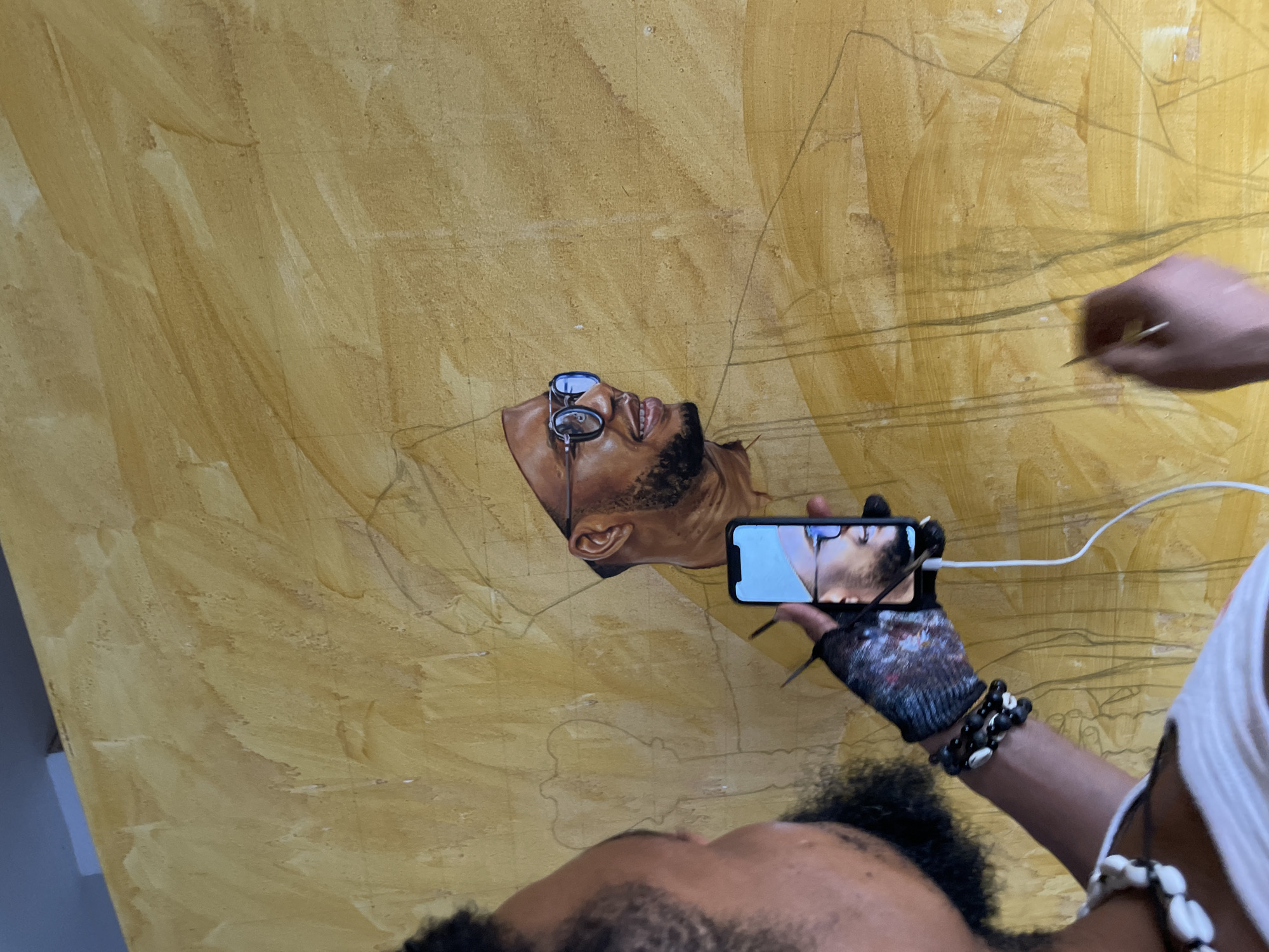 A man in Ado, Nigeria, is painting a portrait on a large, yellow-toned canvas. The portrait depicts a Black man wearing glasses, looking upwards. The artist is holding a smartphone, which displays the same image as the portrait, presumably as a reference. The phone is connected to a charging cable. The artist is wearing a glove on his left hand and beaded bracelets on both wrists. A second hand, holding a paintbrush, is visible on the right side of the image, suggesting a collaborative effort. The canvas shows preliminary sketch lines for the overall composition.