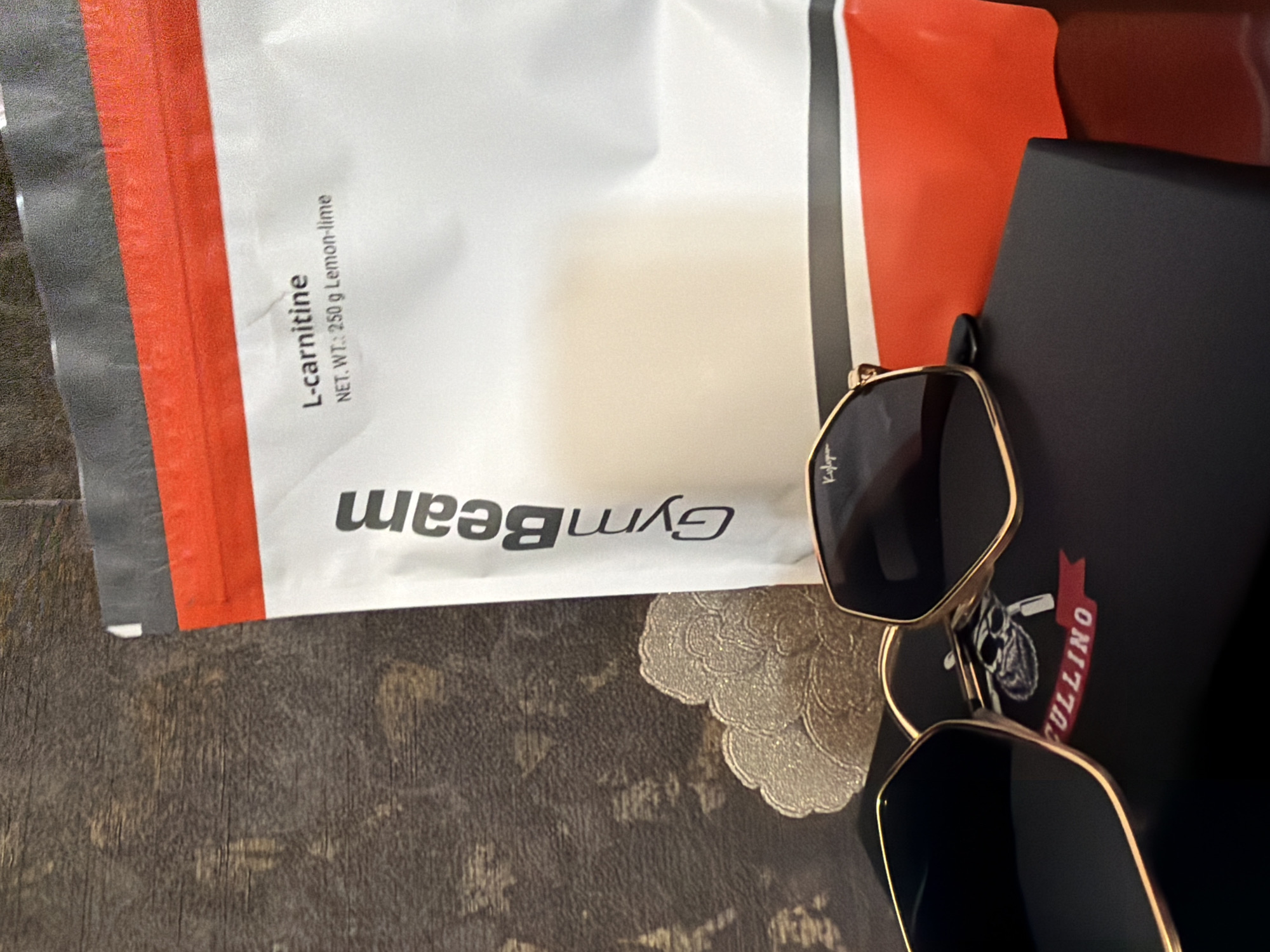 The image shows a GymBeam L-carnitine package (net weight: 250 g Lemon-lime) next to a pair of gold-rimmed sunglasses and a black case with "CULLINO" printed on it. The items are placed on a surface with a floral doily. The location is unknown; no features indicate it is Brusen, Bulgaria.