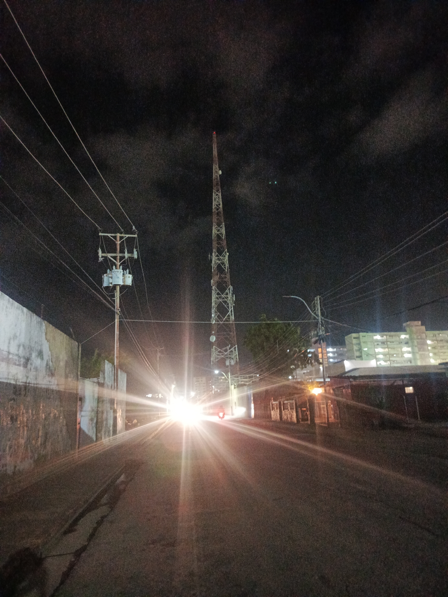 This nighttime street scene in Juan Griego, Venezuela, depicts a paved road extending into the distance under a dark, cloud-laden sky. The primary light source emanates from the center of the frame, manifesting as an intense, starburst-effect glare, consistent with vehicle headlights approaching the viewer. To the right of this central light, a smaller red light is visible, likely a taillight.
On the left side of the road, a weathered, light-colored wall with visible stains and what appears to be faint graffiti stretches into the background. Above this wall, a utility pole is prominent, supporting multiple power lines that extend diagonally across the upper left quadrant of the frame. Three cylindrical transformers are mounted on this pole.
On the right side of the road, a tall, metal lattice communication tower with a red beacon light at its apex dominates the upper right portion of the image. Further down the road on the right, various structures are visible, including a multi-story building with numerous illuminated windows in the distant background, and closer, smaller buildings or residences along the roadside. Sparse vegetation is present between the communication tower and the distant buildings. The road surface itself is dark, showing signs of wear and crack patching. The overall environment is urban or semi-urban, characterized by infrastructure and distant building lights, captured with a grainy texture indicative of low-light conditions. No individuals are discernibly present.