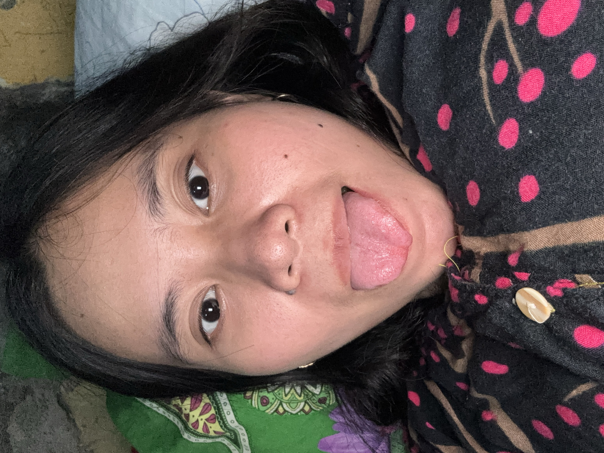 A close-up, selfie-style shot features a young woman lying down with her tongue sticking out. She has dark hair and is wearing a dark top with a pink polka dot pattern and a tan stripe. The background is a mix of green and yellow patterned fabric and a light blue pillow. The image appears to have been taken indoors, likely in a bedroom. No specific location in East Jakarta, Indonesia, can be identified from the visual information provided.