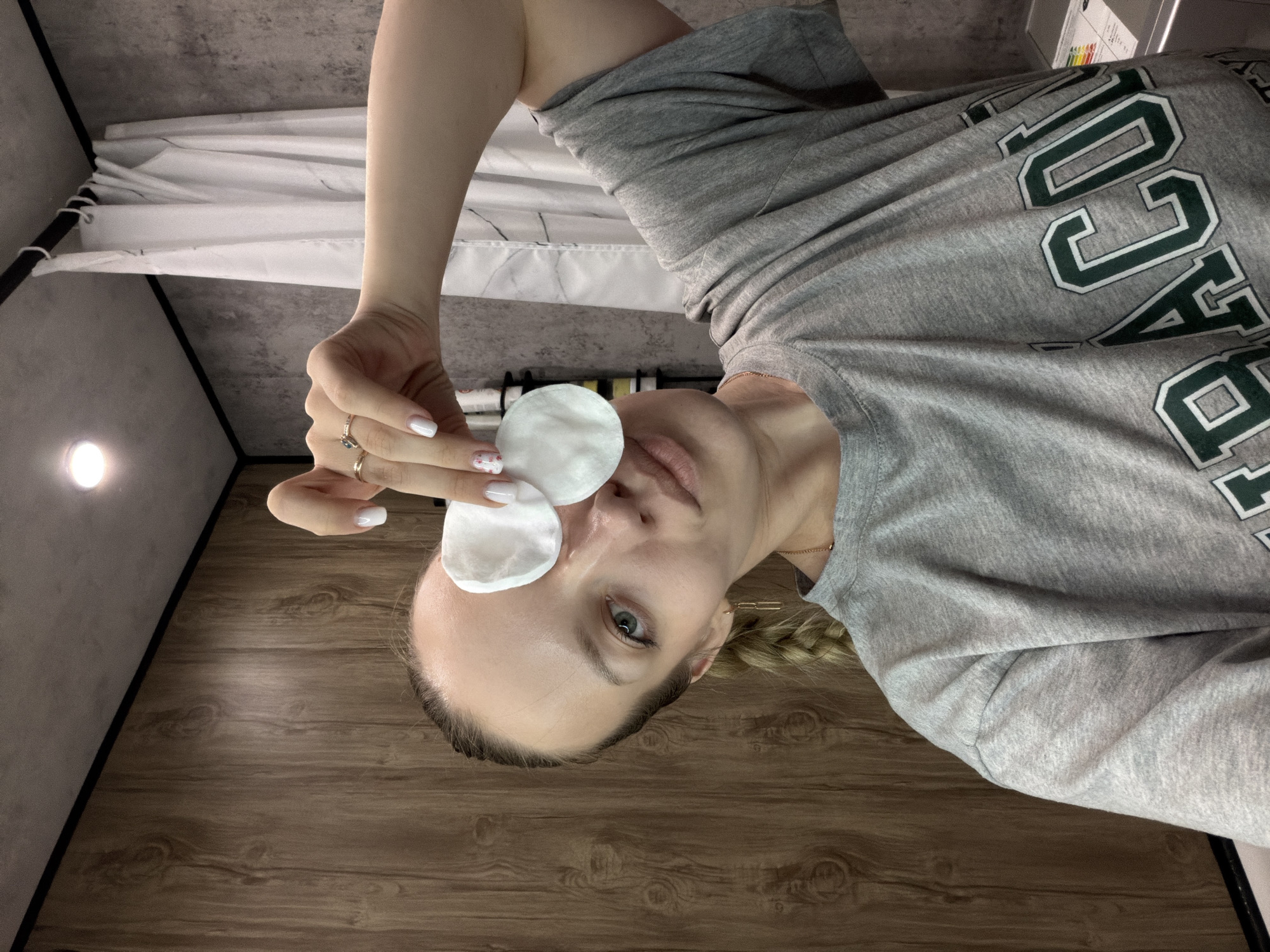A female individual is depicted in what appears to be a residential bathroom in Reshetnikovo, Russia. She has light skin, light-colored eyes, and light hair styled in a braid visible on the right side of her head. She is wearing a grey short-sleeved t-shirt with partially visible dark green and white collegiate-style lettering. Accessories include a gold-colored chain necklace, a geometric gold-colored earring on her right ear, and two rings on her right hand—one a simple gold band, the other featuring a dark stone. Her fingernails are painted white, with one nail displaying red and pink floral detailing. The individual holds two white, circular cotton pads with her right hand, positioning them over her left eye, and is looking directly at the camera with a slight pout. The background on the left consists of vertically oriented wood-grain paneling. The background on the right features a grey, textured wall, possibly concrete or stone-like, from which a white shower curtain hangs on a black rod. A single circular recessed ceiling light fixture is visible in the upper left. A metallic appliance, possibly a washing machine, with a visible energy efficiency label, is partially seen in the lower right corner of the frame.