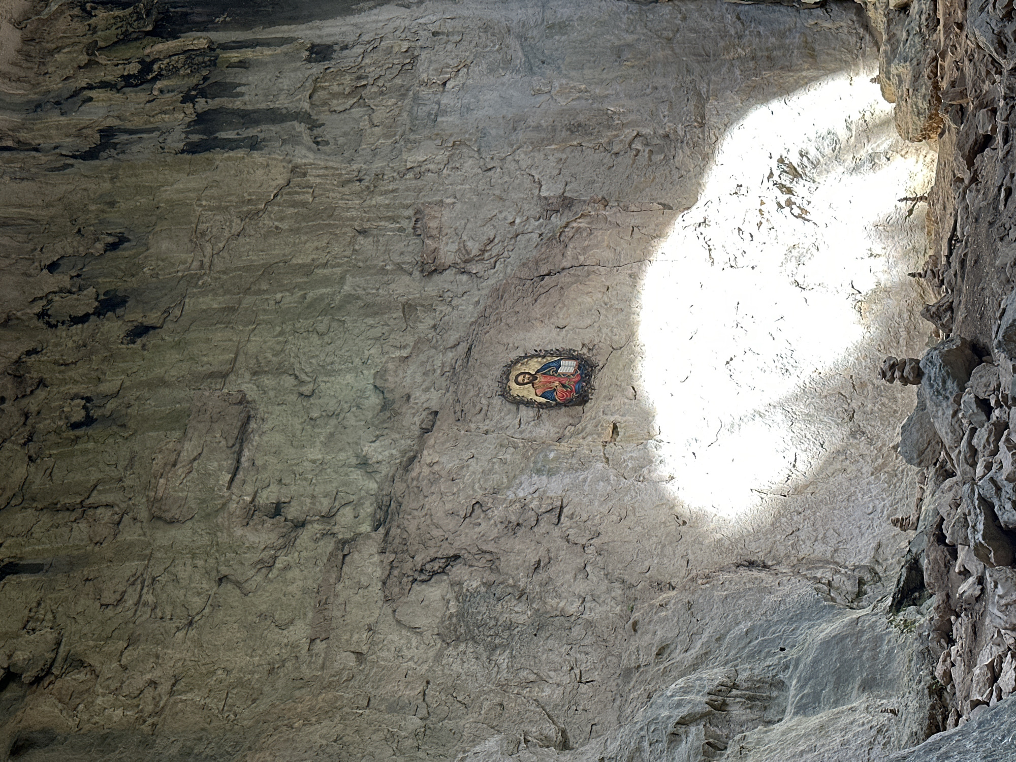 The image depicts a wall inside a cave, possibly in the city of Karlukovo, Bulgaria. The wall is made of rough, grey rock with various cracks and crevices. There's a religious icon affixed to the wall, featuring a depiction of a seated figure with a book in its lap. A strong beam of sunlight illuminates a significant portion of the wall to the right of the icon. A more rugged, rocky formation is visible on the extreme right side of the image.