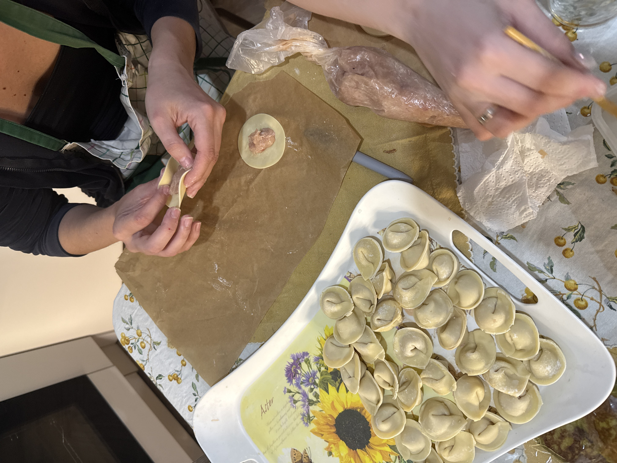 The image shows a person in the process of making dumplings. The person is wearing an apron and has their hands occupied with folding and sealing the dough around a filling. A tray containing a number of already-made dumplings is on the table. Also visible are a bag of filling and a piece of brown paper on which the dumplings are being prepared. There is also an open package. The location might be Krasnoyarsk, Russia, but this cannot be verified with the image alone.