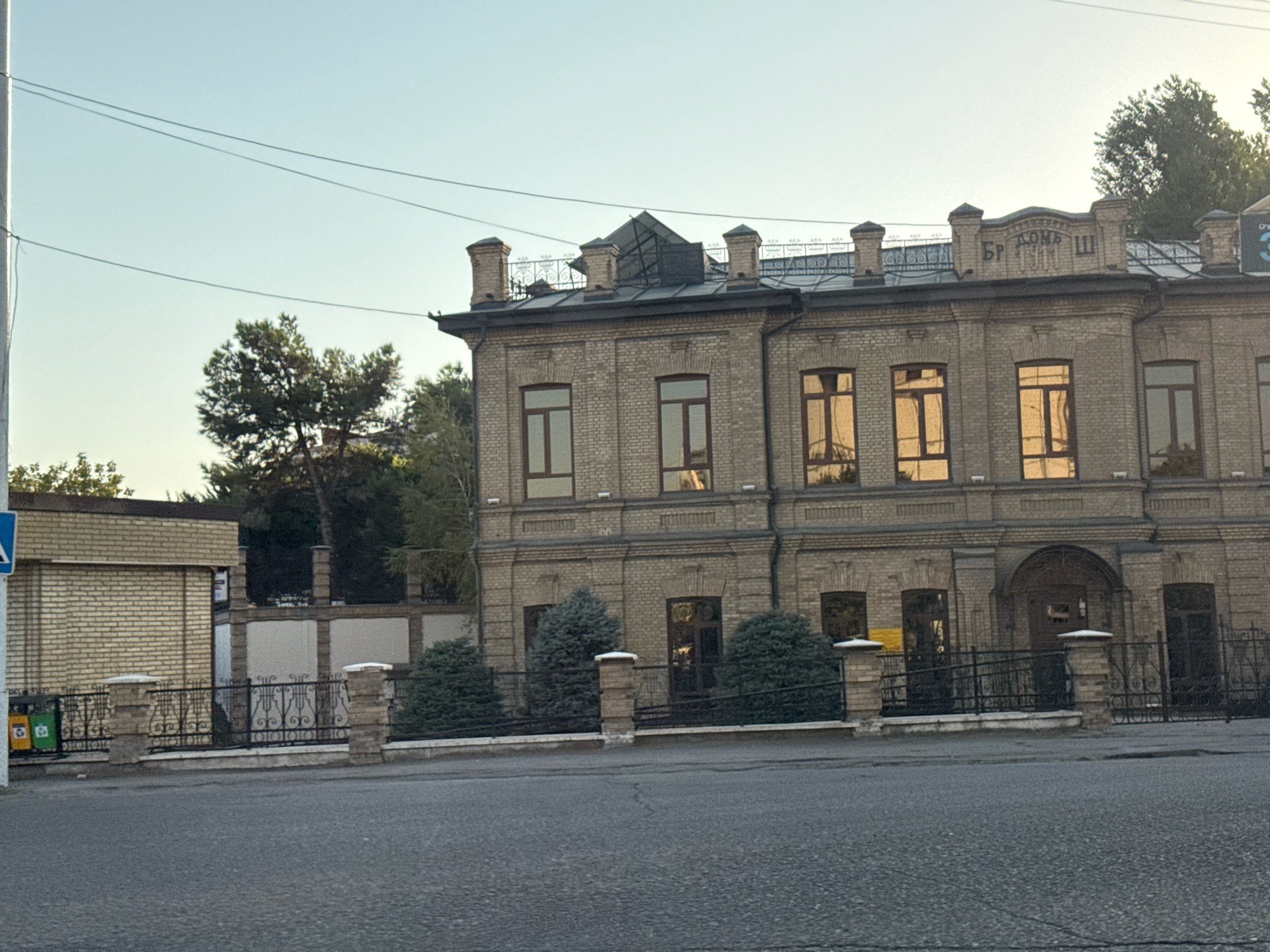 The image shows a two-story brick building with a pitched roof, likely a historic structure, located in Tashkent, Uzbekistan. The building has numerous windows, some reflecting the sky and surrounding trees. A decorative metal fence runs along the front of the property, with brick pillars. To the left, a smaller brick structure is visible, possibly a part of the same complex or a neighboring building. Trees and bushes are present on the grounds, and a street with pavement is in the foreground. Overhead, power lines are strung across the sky. The scene appears to be an exterior shot during daylight, with clear sky overhead.