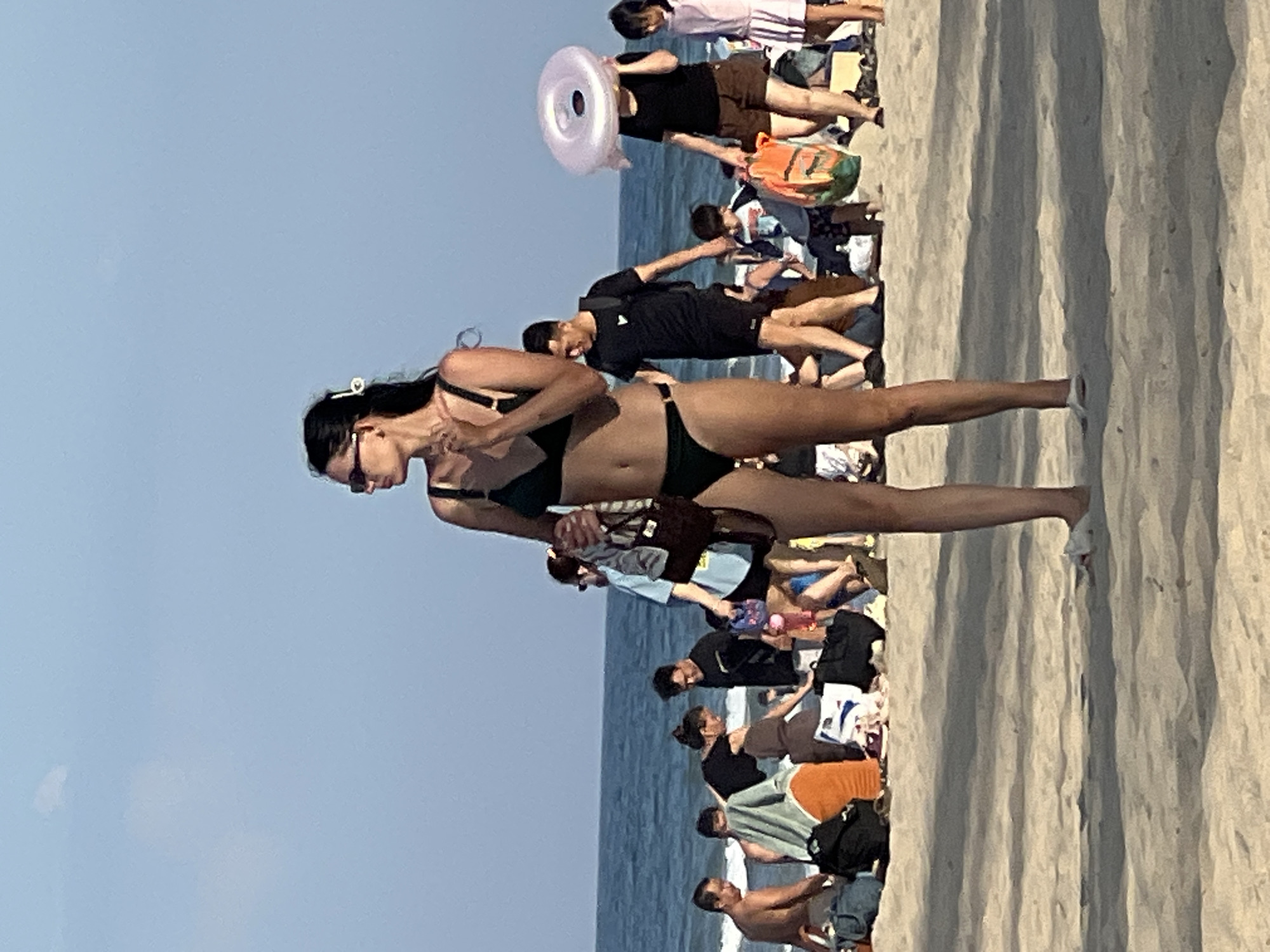 A full-body shot captures a woman in a dark green bikini standing on a sandy beach, looking to her left, holding a dark brown bag in her right hand and a light colored object in her left hand. She is wearing sunglasses and has a white hair clip. The beach is populated with other individuals, some in swimwear, others in casual beach attire, and various beach items like bags, towels, and a child's inflatable ring. The sea and a clear blue sky form the background. The scene is likely My Khe Beach in Da Nang, Vietnam, known for its long stretches of sand and recreational activities.