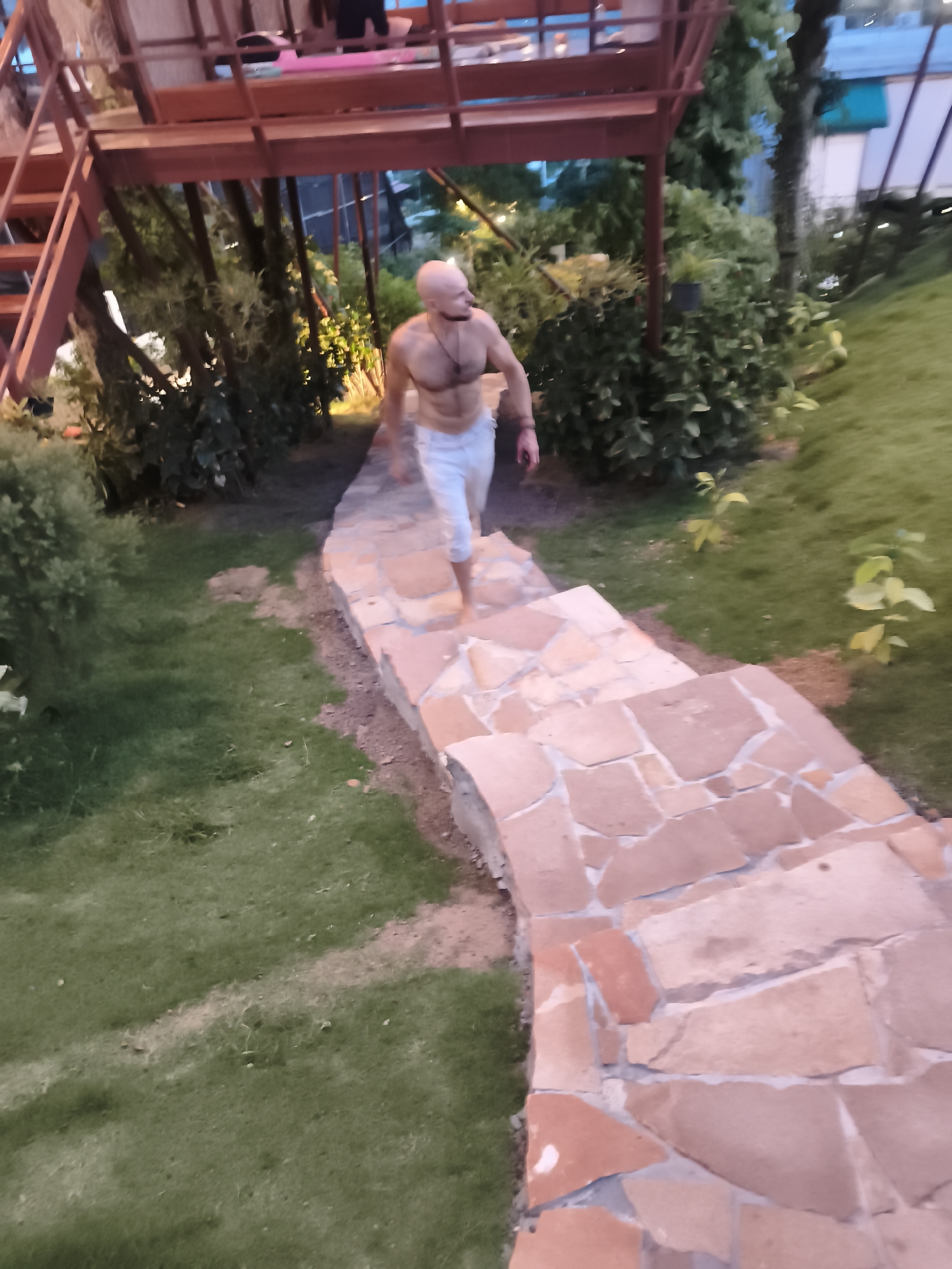 A bald, muscular man with a beard, wearing light-colored pants, walks down a stone pathway. The pathway is bordered by green grass and various green plants. In the background, a multi-level wooden structure with a red railing is visible. The scene appears to be outdoors, possibly in a garden or resort area, during twilight or evening. The location is identified as Ban Bang Rak, Thailand.