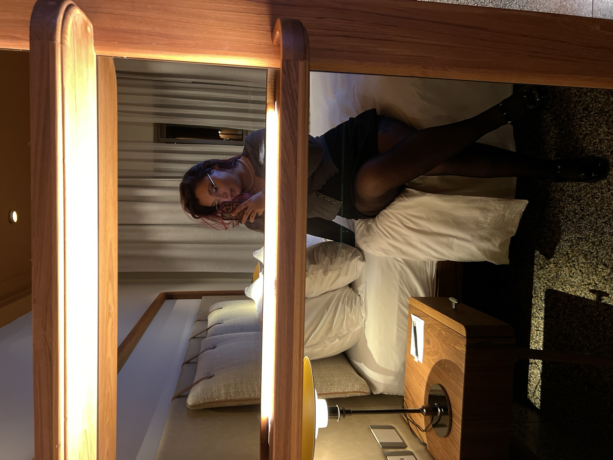 A young woman in black stockings and shoes lies on a bed in a room. The woman is taking a picture of herself with her phone. The room features a unique bed design with a wooden frame surrounding the bed, and a bedside table with a lamp. The location is not specified as being in Tel-Aviv, Israel.