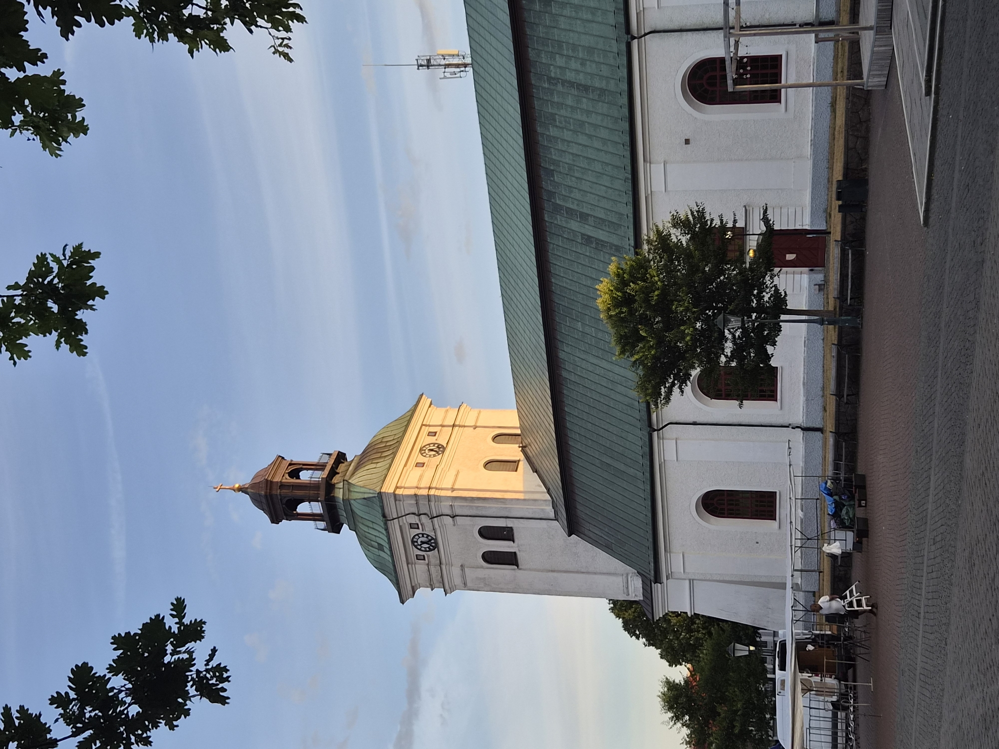 In the city of Varberg, Sweden, a bright white church with a green roof stands prominently under a blue sky with scattered clouds. The church features a tall clock tower with a copper-colored spire topped by a gold cross. Arched windows with dark red frames punctuate the facade of the building. In front of the church, a paved square is set up with market stalls, including a white van and various tables and structures. A man in a white shirt and dark shorts is visible near the stalls, appearing to be interacting with the setup. To the right of the church, a tree with vibrant green foliage grows near the building. Dark green oak leaves frame the top edges of the image, suggesting a natural setting around the urban environment. The overall scene depicts a peaceful daytime setting in a Swedish city square.