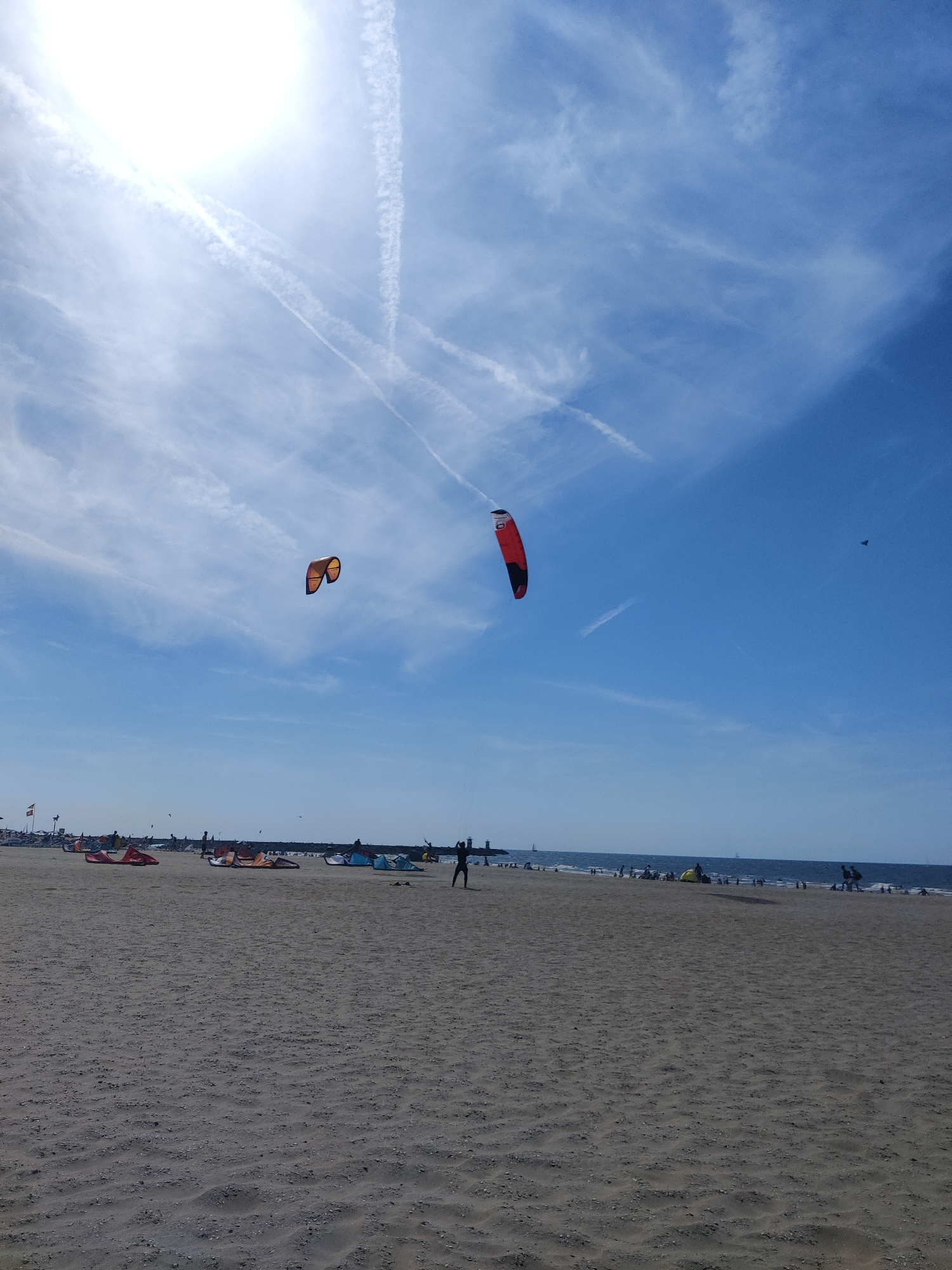 The image depicts a wide, sandy beach on a sunny day with a bright blue sky filled with wispy clouds and contrails. Two kites, one orange and yellow, and another red and black, are visible in the sky. In the distance, a person is kite surfing. The beach is populated with several other kites laid out on the sand, some with people standing near them. Further down the beach, a line of people and structures are visible along the shoreline. The scene appears to be Scheveningen beach in The Hague, Netherlands, given the context provided.