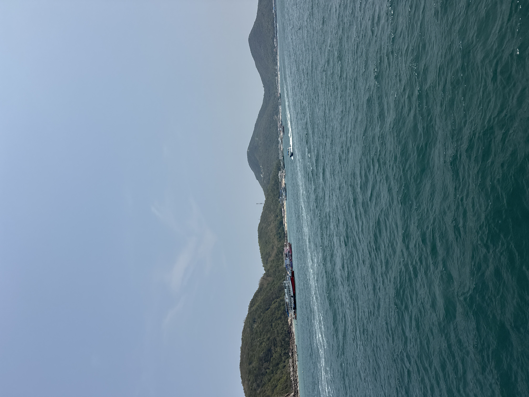 This media file captures a picturesque coastal scene in Pattaya City, Thailand, under bright daytime conditions. The upper portion of the frame features a vast expanse of clear, light blue sky, with only subtle traces of white clouds. Below the sky, running horizontally across the image, is a verdant coastline composed of undulating hills and mountains densely covered in green foliage.
The lower and right sections of the image are dominated by an expansive body of deep blue-green water, likely the sea or ocean. The water's surface is textured with numerous ripples and gentle waves, reflecting the sunlight and suggesting a calm yet active environment.
Along the coastline, several boats and vessels are visible. In the mid-ground, a white speedboat is actively moving across the water, leaving a distinct white wake behind it. Closer to the foreground on the left, a cluster of larger vessels, possibly passenger ferries or tour boats, are docked or moored near the shore. One prominent boat among them features a noticeable red hull. Small buildings or structures can be discerned intermittently along the edge of the land.
The overall impression is one of peaceful maritime activity and natural beauty on a sunny day. There is no visible text in the image.