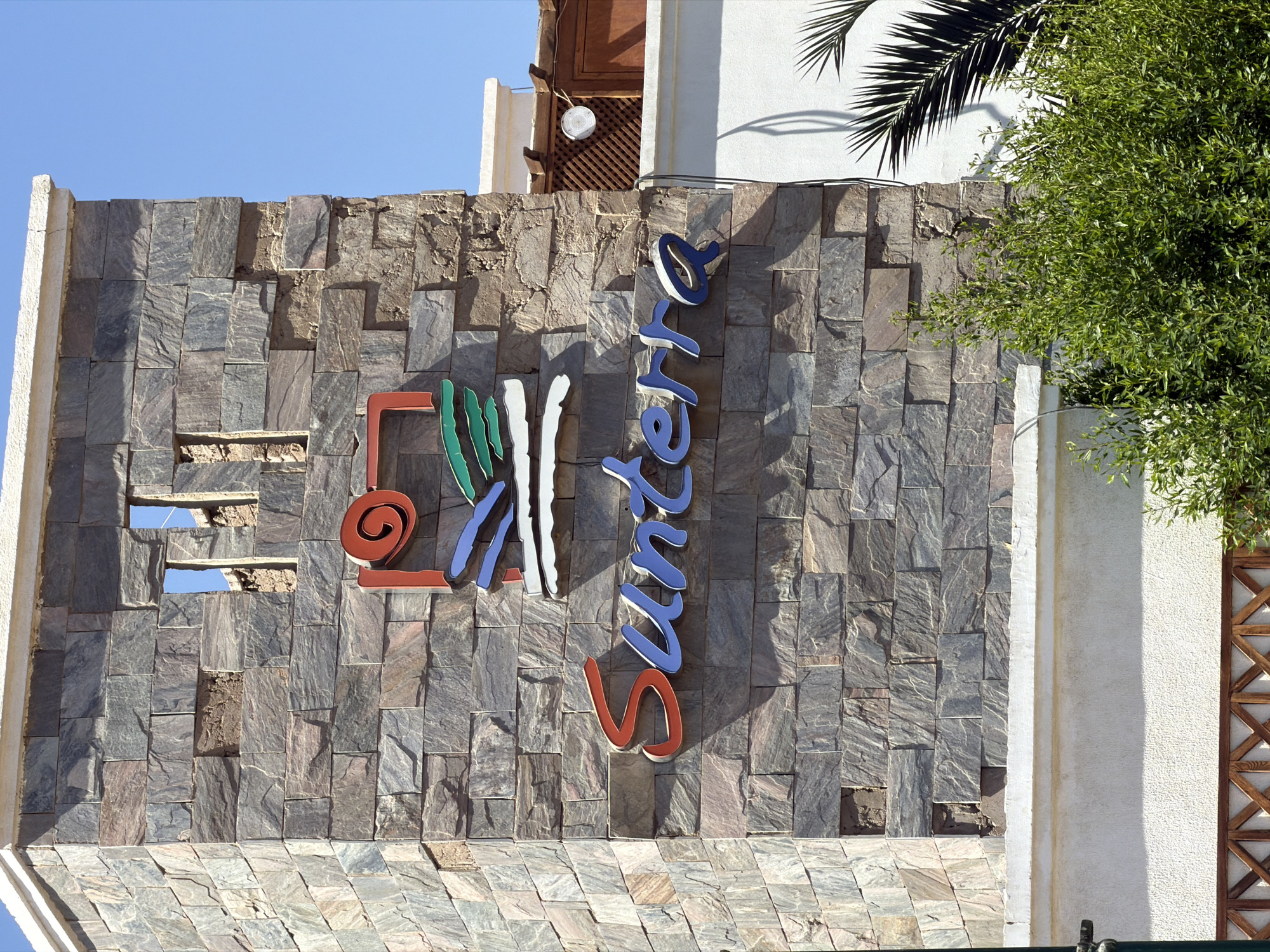 A stone-clad wall with a blue and white sign reads "Santerra" vertically. To the left of the sign, a graphic includes a red spiral shape and green, white, and blue vertical lines. The wall is textured with irregularly shaped stone tiles in shades of gray and brown. Above the wall, part of a white building with wooden lattice and a palm frond are visible against a clear blue sky. The scene suggests a sunny day, likely in a warm climate. The stonework appears somewhat weathered, with some missing tiles. The overall impression is of an exterior facade, possibly at a resort or business in Sharm El Sheikh, Egypt. No people are visible in the image.