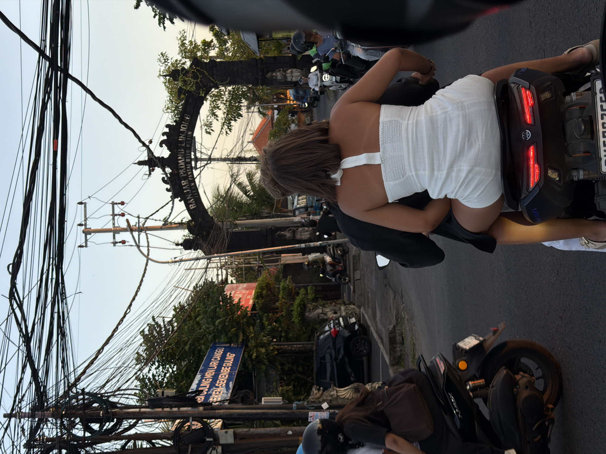 A rear-view, medium shot captures a couple riding on a scooter in North Kuta, Indonesia. The woman in front wears a white strapless romper and the man behind her is dressed in a black jacket. They are on a street with numerous overhead power lines and a large stone archway that reads "BANJAR PADANG LINJONG" and "JALAN PANTAI BATU MEJAN." In the background, other scooters, a black car, and a few buildings are visible. The sky is overcast.