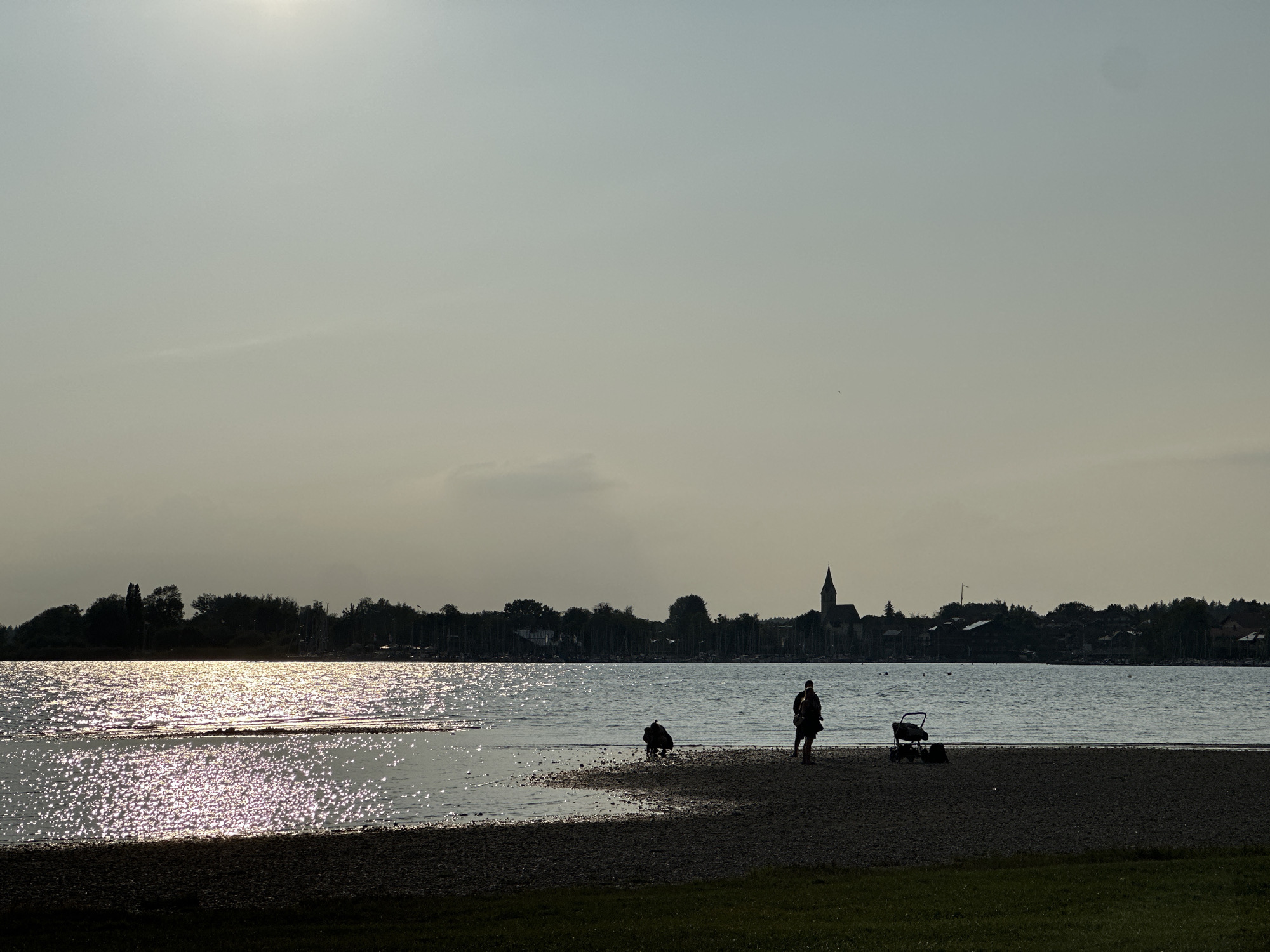 In the city of Chieming, Germany, a tranquil lakeside scene unfolds as the sun casts a shimmering golden glow across the water. Silhouetted figures dot the shoreline. One person stands, observing the water, while another kneels, possibly interacting with the water or sand. A stroller sits nearby, implying a family outing. The background is a soft blur of trees and buildings, with a prominent church steeple rising above the treeline. The sky is a pale, hazy expanse, suggesting either early morning or late afternoon light. The image evokes a sense of calm and peacefulness, capturing a moment of quiet enjoyment by the lake.