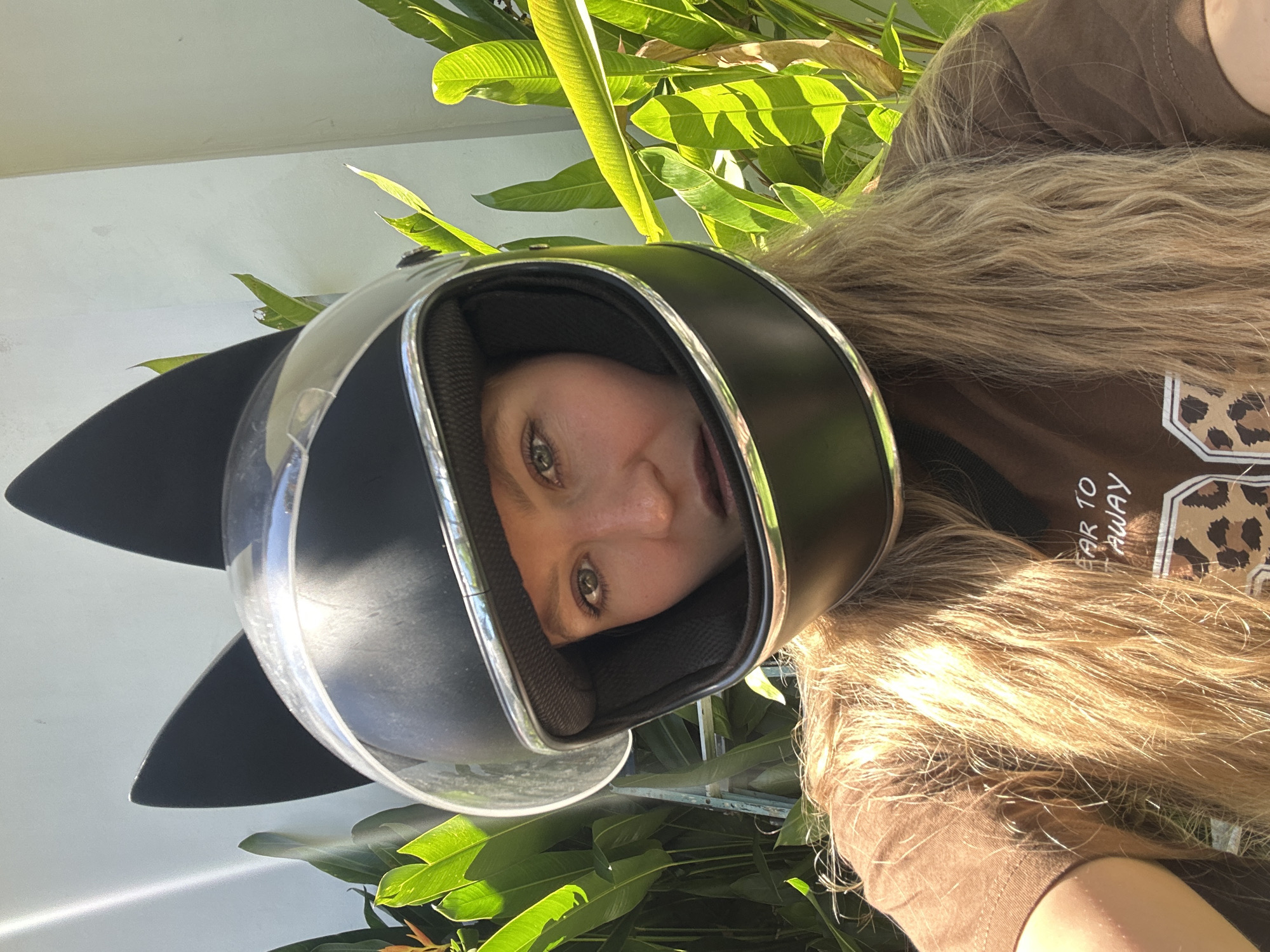 The image presents a close-up, portrait-oriented shot of a person wearing a distinctive motorcycle helmet, set outdoors against lush greenery and a light-colored wall. The original image is rotated 90 degrees counter-clockwise.
The subject is a person, likely female, with long, wavy blonde hair that falls over her shoulders. She is wearing a black full-face motorcycle helmet, characterized by pointed, cat-ear-like attachments on the top and a clear visor. Her face, framed by the helmet's opening, is visible. She has light-colored eyes (possibly green or hazel) and a slight smile or playful expression, looking directly at the camera.
She is dressed in a short-sleeved brown top, which features a graphic design incorporating a leopard print pattern. Visible text on the top reads "EAR TO AWAY".
The background consists of vibrant green plant foliage, brightly illuminated by natural sunlight, suggesting a tropical outdoor environment. A plain, light-colored wall is also visible behind her. The strong sunlight indicates a clear, sunny daytime, casting distinct highlights and shadows.
The scene depicts a casual moment, with the person posing for a photo. The tropical foliage aligns with the given location context of Kuta Selatan, Indonesia.