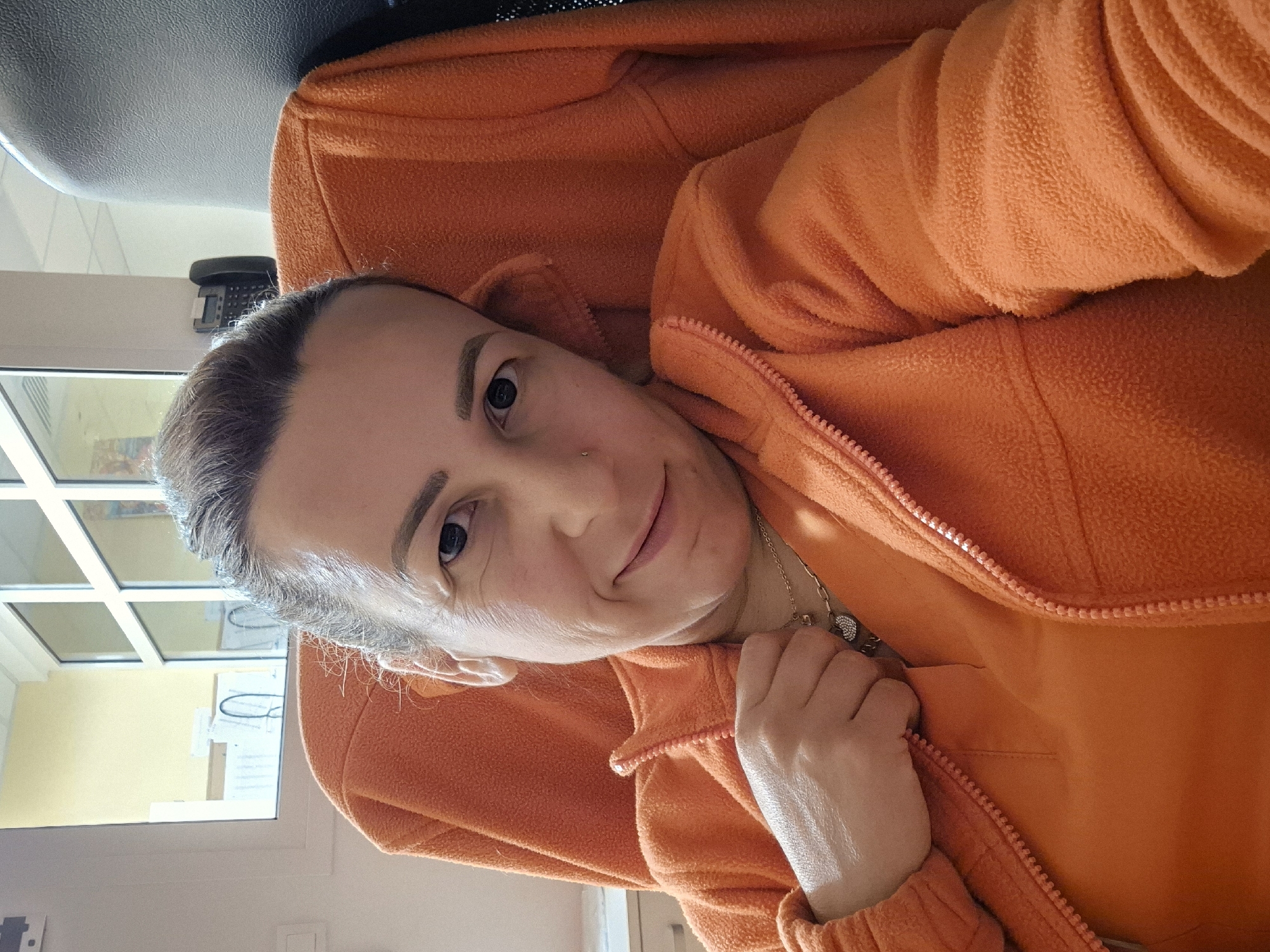 A woman with blonde hair pulled back and wearing an orange fleece jacket smiles at the camera in a selfie. She is seated in a chair, and a portion of a dark gray chair is visible to her right. Behind her, a window with white frames looks out onto a room with yellow walls, suggesting an indoor setting, possibly a hospital or clinic given the presence of medical equipment and a landline telephone mounted on the wall. The woman is wearing a silver necklace with a pendant. The image is taken from a close-up perspective, focusing on the woman's face and upper body.