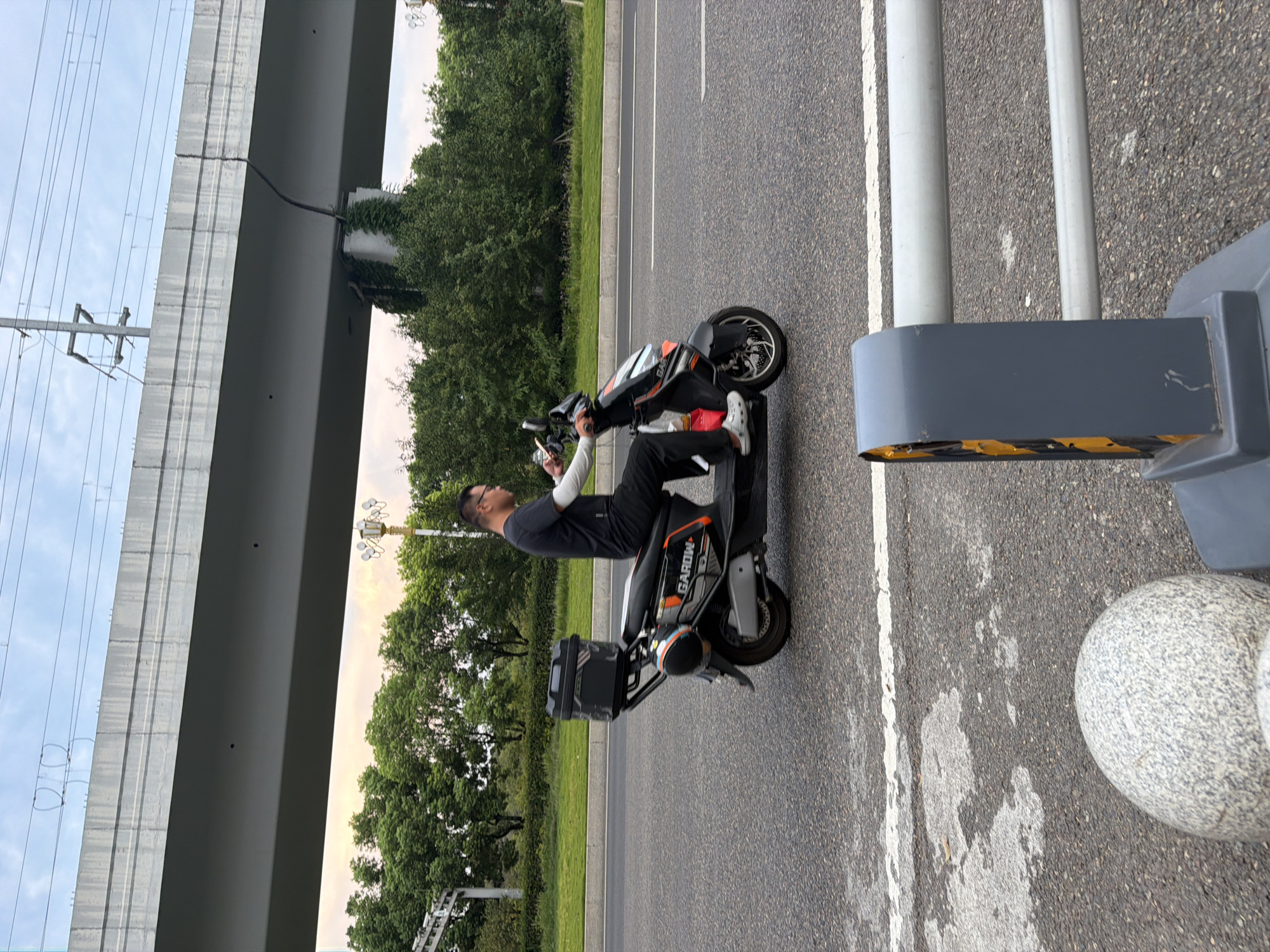 A man is riding a black and orange electric scooter on a paved road in Jiangjin, China. The scooter has a black storage box mounted on the rear. The man is wearing a dark t-shirt, black pants, and white Crocs. He is looking forward and holding the handlebars. The road has white lane markings and appears to be a multi-lane road. In the background, there are trees, a grassy median, and an elevated concrete structure, possibly an overpass or bridge, with electrical wires running above it. The sky is overcast with hints of the sunset. In the foreground, there is a gray bollard with yellow and black striped warning tape.