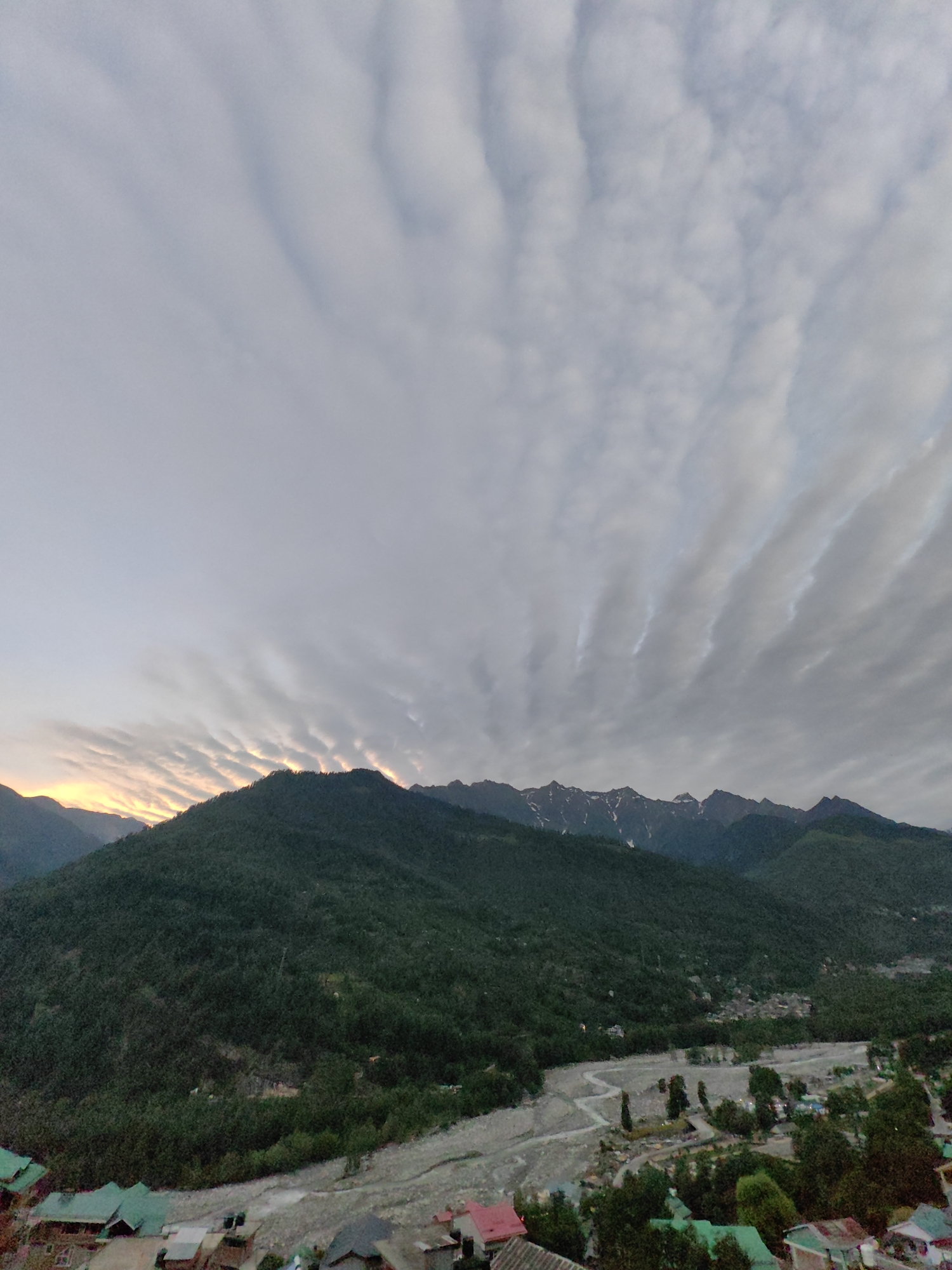 The image captures a wide-angle, elevated view of a valley in Vashisht, India, at dusk. The sky is dominated by a dramatic display of altocumulus undulatus clouds, characterized by parallel wave-like formations, tinged with the soft glow of the setting sun on the left horizon. Below the sky, a range of lush, green mountains stretches across the frame. The slopes are densely covered with trees, and in the higher altitudes, snow-capped peaks are visible. A riverbed, appearing dry or with very shallow water, winds its way through the valley floor. Scattered along the river and nestled at the base of the mountains are numerous small buildings with distinctly colored roofs, primarily green and red, suggesting a village or small town. The architecture appears to be traditional. The overall scene is serene and highlights the natural beauty of the mountainous landscape and the human settlement within it.