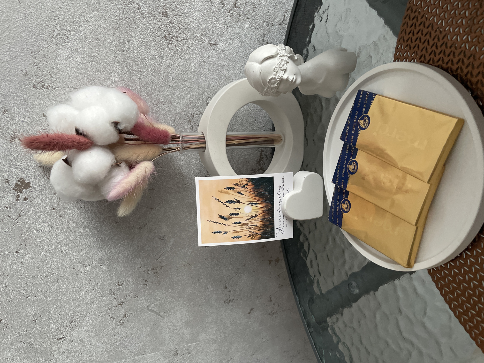 A top-down shot shows a table with a gray surface. On the table are several decorative items and treats. To the left is a bouquet of dried flowers in a ring-shaped vase attached to a statue of an angel. Next to the vase is a rectangular card with an image of wheat stalks set against a sunset and a white heart-shaped object. On a small white plate are three golden-wrapped Merci chocolate bars, their blue wrappers slightly visible at the edges. The table extends to the right, where a corner of a brown woven placemat can be seen. The image does not provide specific information to locate the scene in the city of Izhevsk, Russia.