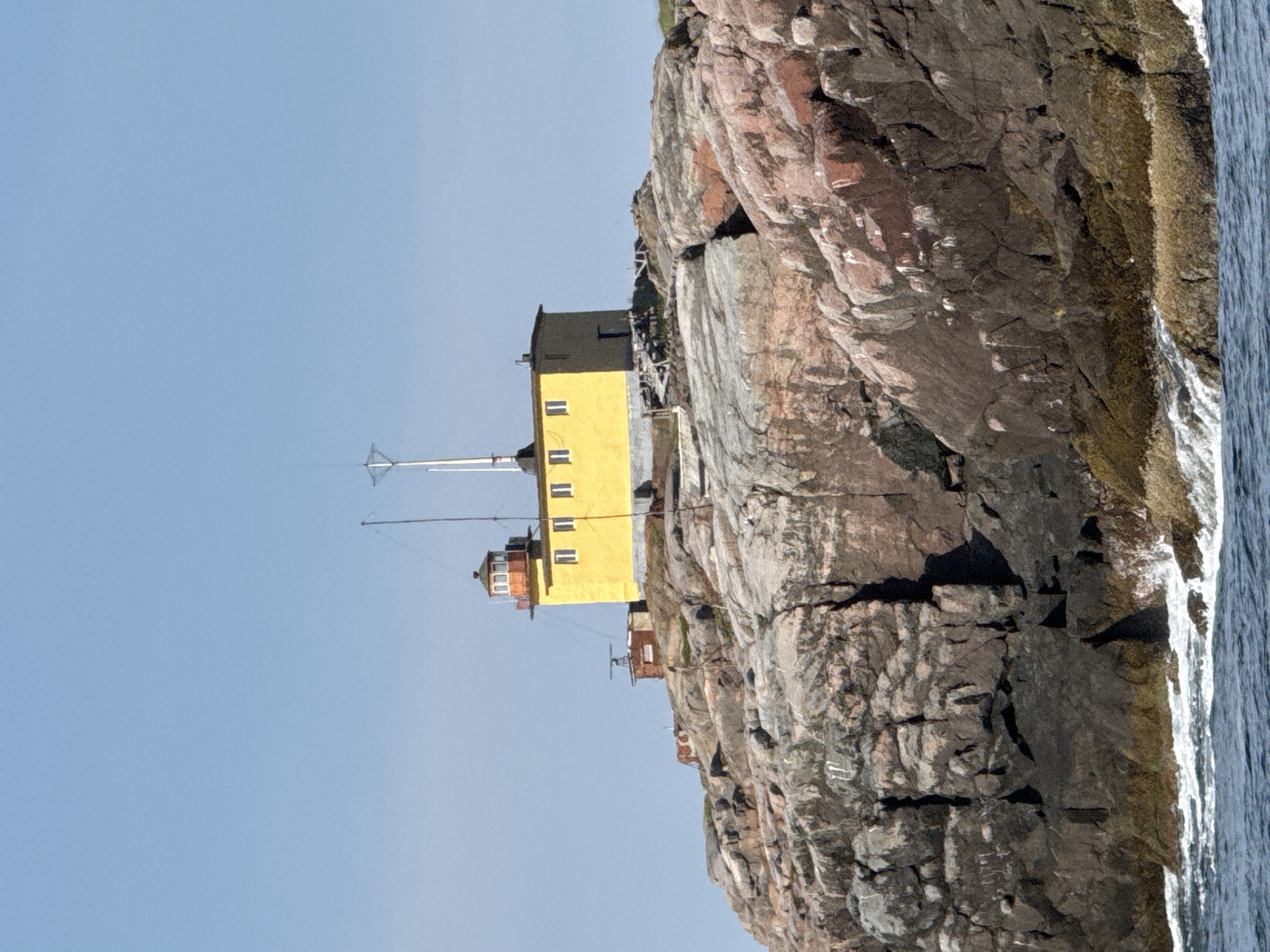 This image shows a remote lighthouse situated on a rocky outcrop in Teriberka, Russia. The lighthouse, painted in bright yellow, has a red-topped lantern room. Adjacent to the lighthouse are smaller, brick structures and a metallic antenna. The rocky terrain surrounding the lighthouse is rugged and appears to be a natural formation. The scene is set against a clear blue sky, with the ocean visible at the bottom of the frame, showing waves lapping against the rocks. There are no people visible in the image. The overall impression is one of isolation and a harsh, natural environment.