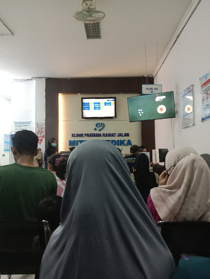The image displays an interior view of a medical clinic's waiting area in Gladaksari, Indonesia, identified by signage as "KLINIK PRATAMA RAWAT JALAN MITRA MEDIKA" (First-Level Outpatient Clinic Mitra Medika). The perspective is from behind several seated individuals, looking towards a reception or information counter.
In the foreground, the backs of at least three people are visible: one individual in a dark green t-shirt on the left, another wearing a dark grey full-body covering in the center (with a child's head partially visible below), and a person in a light pink/beige hijab on the right, turned slightly to their right. They are seated on black-framed chairs.
The midground features a counter area with a prominent blue-and-white sign reading "KLINIK PRATAMA RAWAT JALAN MITRA MEDIKA." Above this sign, two wall-mounted flat-screen monitors are active. The left monitor displays a blue interface with columnar data, while the right monitor shows a green background with multiple white dots and a larger red circle, likely a queue display. A small white overhead sign points down towards the right monitor, labeled "POJOK JKN" (JKN Corner). Behind the counter, clinic staff are present; one individual is clearly visible wearing a light blue medical mask and a dark uniform.
The environment features white walls and a white ceiling with recessed lighting and a white oscillating ceiling fan. Several informational posters are mounted on the right wall. An informational stand with blue placards is visible on the far left. A security camera is mounted on the ceiling in the upper-right quadrant.