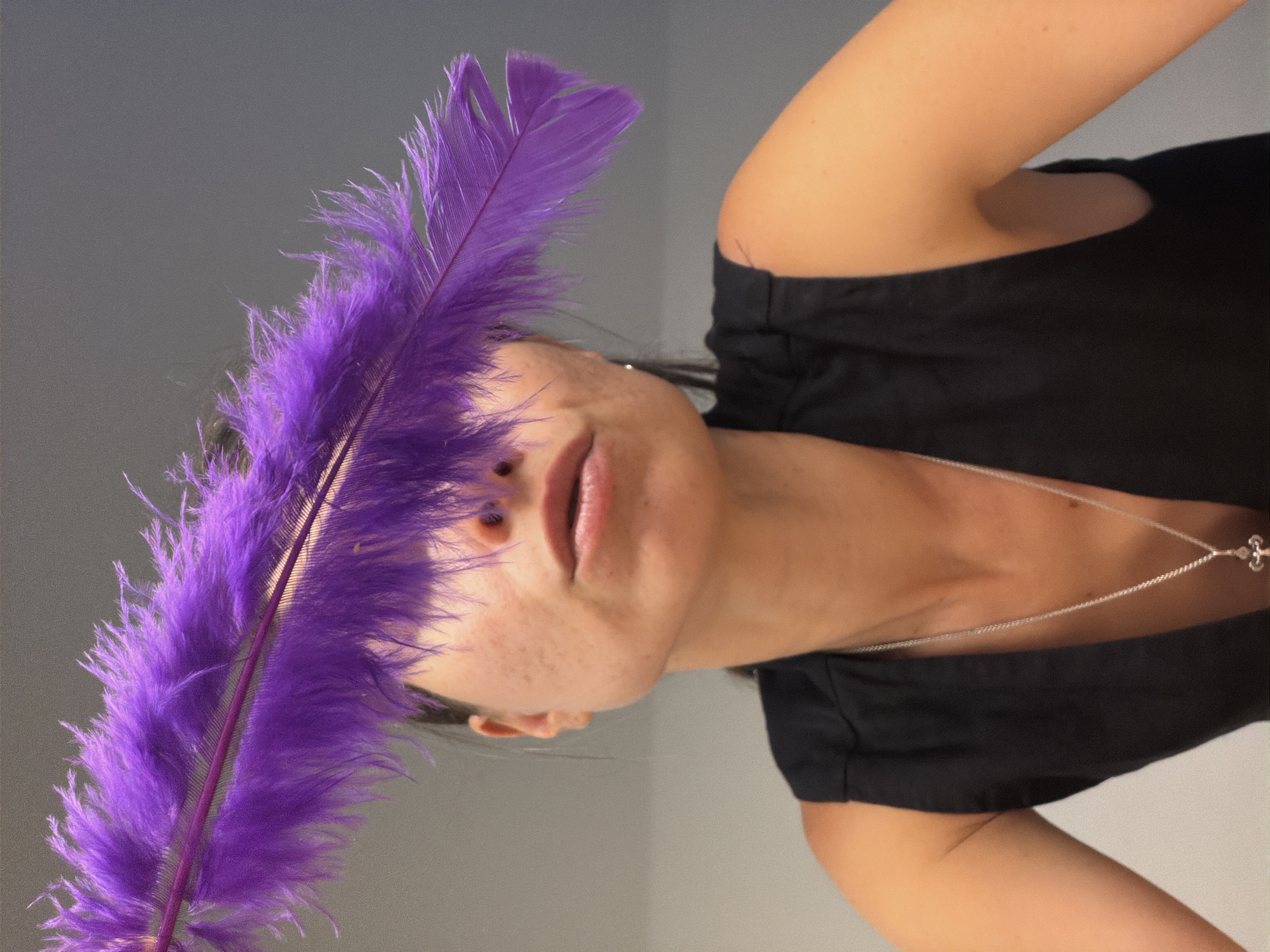An artistic portrait captures a person, likely female, with a large, vibrant purple feather partially obscuring their face. The subject's head is tilted, and their gaze is directed upwards, with soft, parted lips. Visible features include a portion of their chin, cheek, and a smattering of freckles. A delicate silver necklace with a small, indistinct pendant, possibly a cross, adorns their neck.
The person is wearing a simple black top or dress, revealing a bare shoulder and upper arm. The prominent purple feather, with its soft, intricate barbules and a distinct central quill, takes up a significant part of the left side of the frame. Its striking color provides a strong contrast against the person's skin tone and the plain background.
The setting appears to be indoors, against a minimalist light grey wall, which keeps the focus on the subject and the feather. The lighting is soft and diffused, enhancing the textures present. The overall mood is serene and artistic, emphasizing visual composition rather than a specific activity. While the location context is Budva, Montenegro, no specific geographical cues are visible within the image itself. No text is visible.