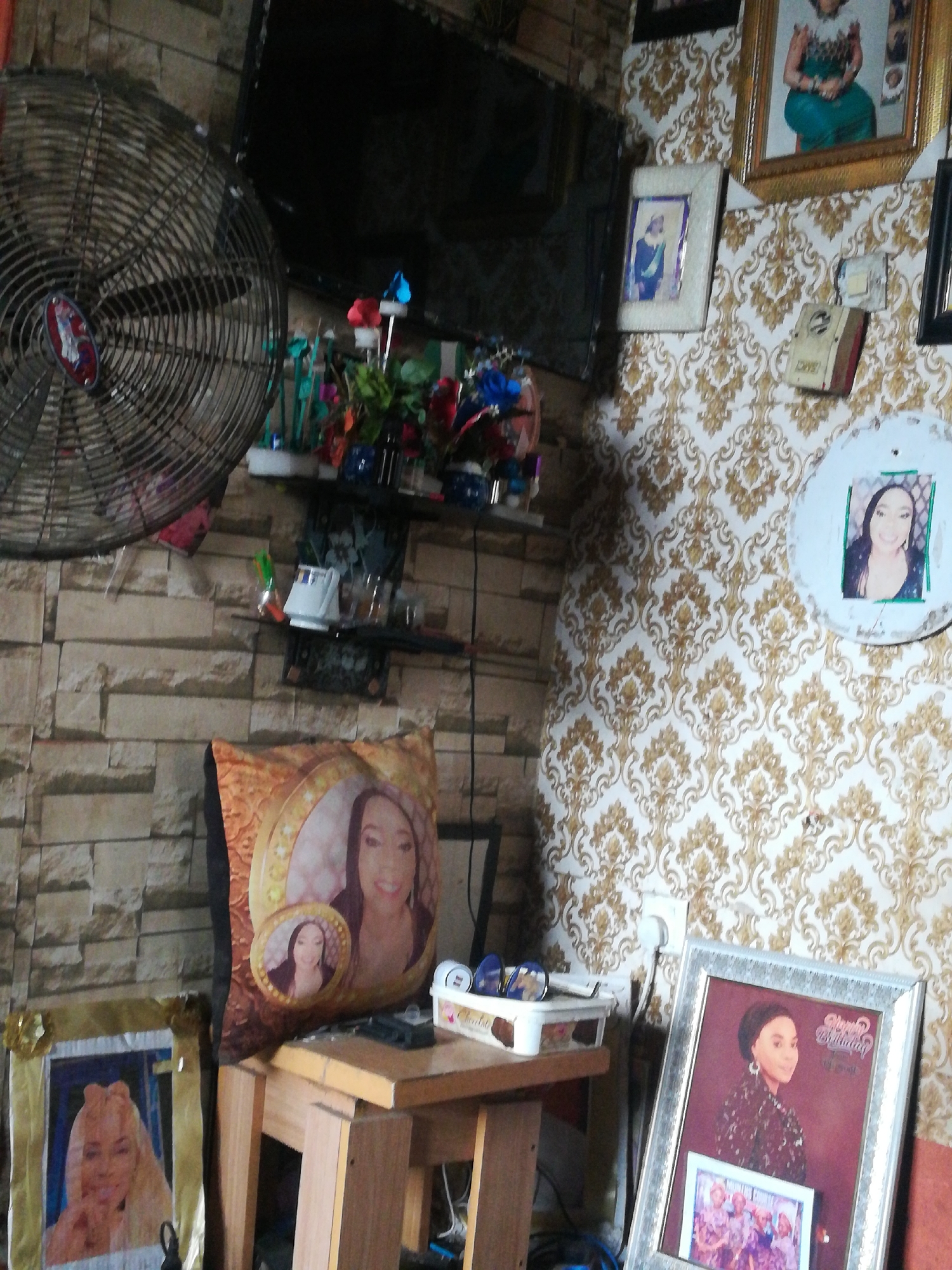 This image captures a densely decorated indoor room, likely a living space, filled with personal items and photographs. The room features two distinct wall patterns: one simulating light-colored brickwork on the left, and another with an ornate white and gold damask wallpaper on the right.
Dominating the upper left is a large, dark wall-mounted fan. Below it, a dark-screened television is mounted above a small shelf displaying various decorative items, including colorful artificial flowers and small figurines.
The room is adorned with numerous framed photographs and printed images, predominantly of women. A large gold-framed picture on the damask wall depicts a woman in a vibrant green dress. Below it, a smaller framed picture shows a man in what appears to be formal attire. Further to the right, a circular white wall hanging features a picture of a smiling woman. An old-fashioned book or box, labeled "LOVE," hangs on the damask wall next to a white electrical outlet.
In the lower-center, a brown wooden stool or small table holds a pillow printed with images of a woman. On this table, sunglasses rest on a white box. Leaning against the table on the right is a large framed picture with the text "Happy Birthday?" and a date "10.01.2001," showing a woman in a patterned top. To the far left, another framed picture on the floor displays a woman with blonde hair.
The overall impression is one of a personalized space, celebrating individuals and possibly family events, with a focus on portraiture. No direct activity is taking place.