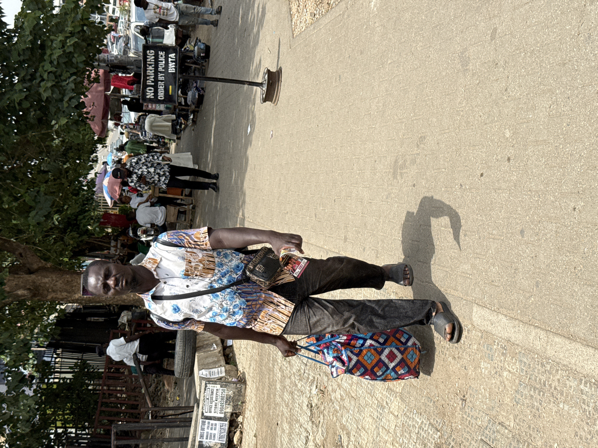 The image depicts a vibrant street scene in Abuja, Nigeria, captured during bright daylight, evidenced by the strong shadows cast on the light-colored paved ground. The primary subject is a man of dark complexion, positioned in the foreground and walking along the pavement. He is dressed in a distinctive short-sleeved shirt featuring white, blue, and brown patterns, dark trousers, and dark open-toed sandals. Across his chest, he wears a black fanny pack, and in his left hand, he carries a colorful bag with a geometric pattern in blue, orange, red, and white. His expression is neutral.
The background reveals a bustling urban environment, likely a market or commercial area. A prominent black sign in the mid-ground reads "NO PARKING ORDER BY POLICE BWTA." Beyond this sign, numerous cars are parked, and many other people are visible, suggesting a crowded street. Some individuals in the distance are seen under umbrellas or makeshift shelters, possibly vendors or people seeking refuge from the sun. To the left of the main subject, a sign advertises "CHRIS E BOTIQUE" and lists job opportunities such as "OFFICE ASSISTANCE" and "STORE KEEPER," along with phone numbers: 07062838755 and 09049380250. Green foliage from trees is visible on the far left, adding a natural element to the urban landscape. The overall scene conveys the active daily life of a busy city street.