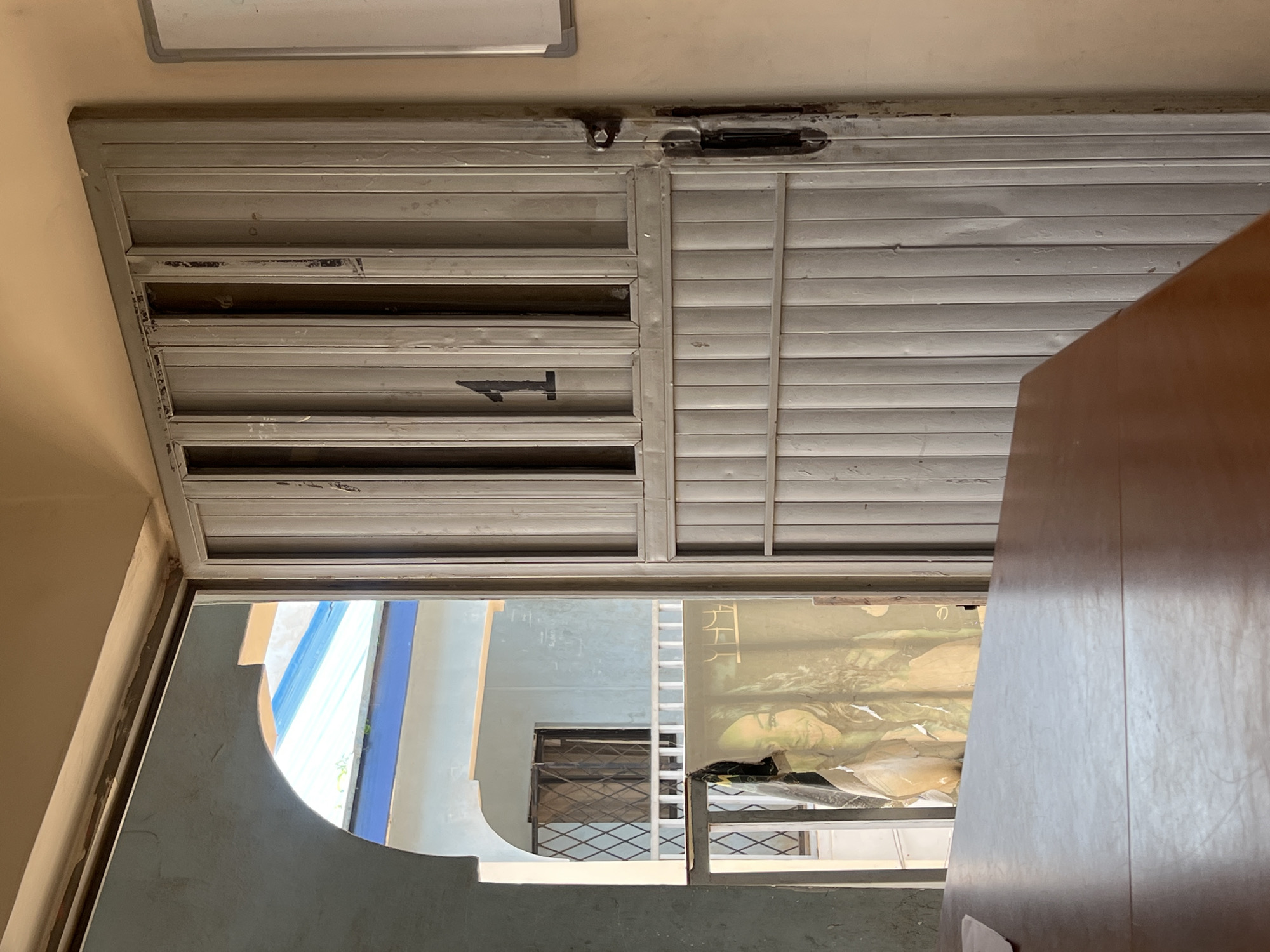The image shows a view through an arched doorway into an interior space with a metal door. The door is gray and has vertical slats with several rectangular openings. A large black number "1" is visible on the upper section of the door. To the left of the door, through an archway, a poster or image of a woman is partially visible, appearing to be tacked to a railing. Beyond the poster, there is a building with a blue and white roof and barred windows. The foreground is dominated by a brown wooden surface, possibly a table. The scene appears to be in Jos, Nigeria, given the context provided.