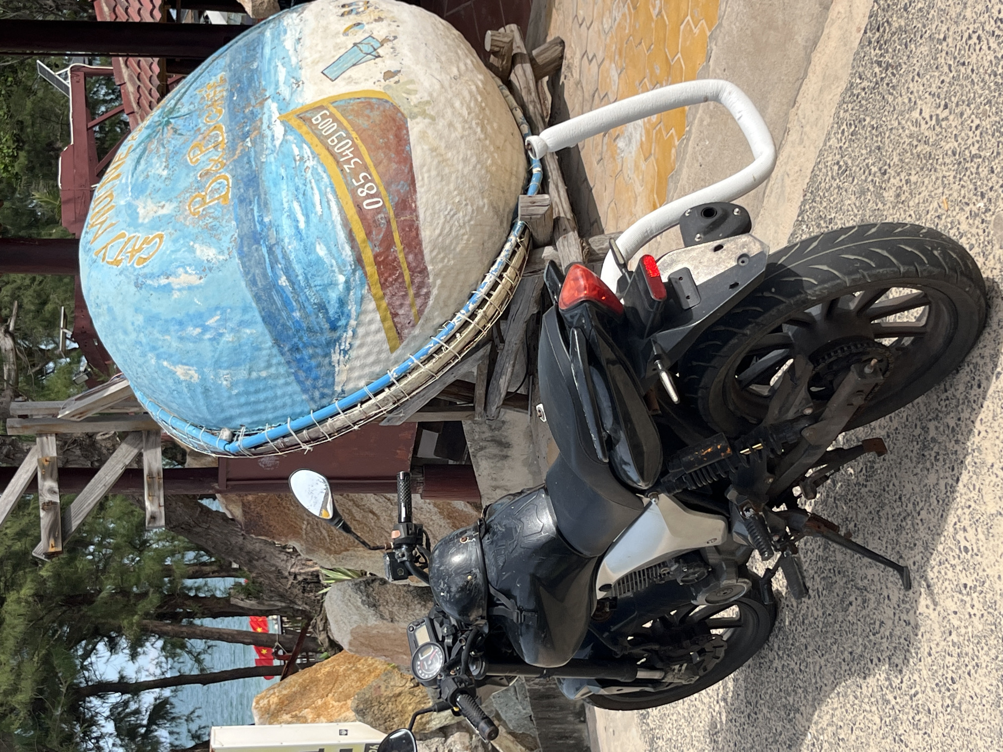 A black motorcycle is parked on a paved surface next to a large, blue, spherical object adorned with a painted beach scene and a phone number. The motorcycle, a naked sportbike model, has a black helmet resting on its fuel tank. Behind the motorcycle and the spherical object, a body of water is visible, suggesting a coastal location. Trees with sparse foliage are present, along with large rock formations. The overall impression is of an outdoor scene, possibly near a beach or a recreational area. The scene is consistent with locations in Phu Thuy Ward, Vietnam, a coastal area known for its beaches and fishing communities.