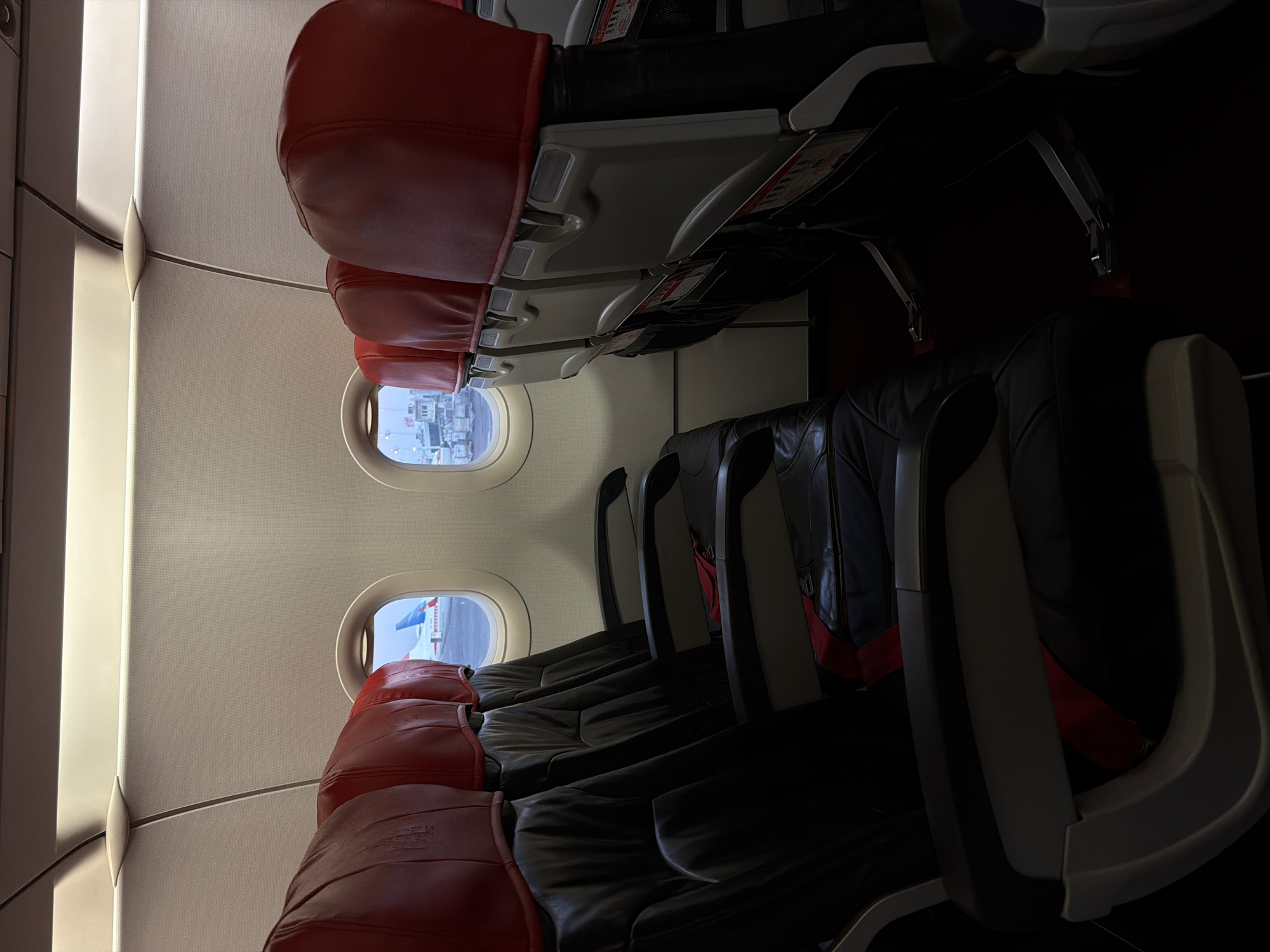 An interior view from an airplane cabin shows rows of empty seats. The seats are upholstered in black leather with red headrests. Two oval windows look out onto an airport tarmac. On the left, another airplane is visible, its tail fin white with blue and red markings. On the right, airport infrastructure with a control tower is visible. The lighting in the cabin is a combination of overhead fluorescent lights and ambient light from the windows. The image is taken from a low angle, looking down the aisle of the aircraft. The location is the Denpasar Airport in Kuta, Indonesia.
