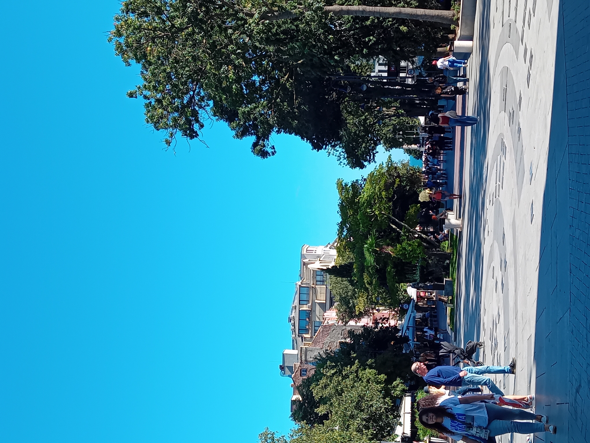 The image depicts a sunny day in Varna, Bulgaria, specifically on the pedestrian promenade near the Sea Garden. The scene is a wide, paved walkway with an intricate geometric pattern.
In the foreground, several people are walking. To the left, a woman in a white t-shirt with graphics and light blue jeans walks towards the camera. Beside her, a man in a blue long-sleeved shirt and blue jeans walks away from the camera, pushing a stroller. Further down the promenade, a group of people strolls in the distance, some of whom appear to be tourists.
The promenade is lined with mature trees on both sides, casting shadows on the pavement. On the left, there are buildings with various architectural styles, including some older, multi-story structures with balconies and windows, and some modern elements like satellite dishes. On the right, the trees are denser, creating a more park-like atmosphere. Beyond the trees on the right, a glimpse of the Black Sea is visible in the distance. Street lamps are also present along the promenade. The sky is a clear, bright blue.