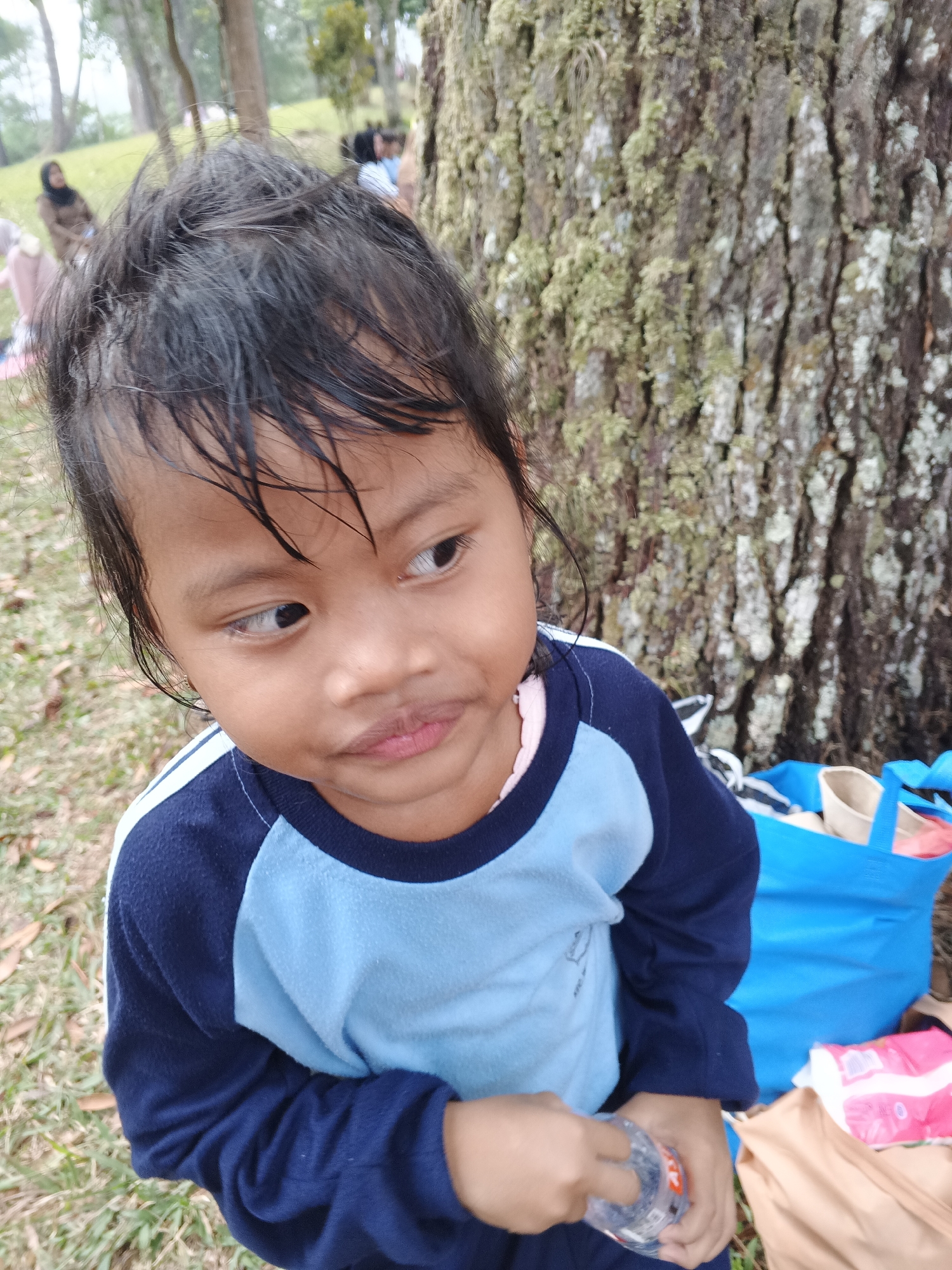 A young child with dark hair and brown skin, wearing a blue and light blue long-sleeved shirt, stands in front of a large tree with textured bark. The child is holding a clear plastic bottle in their right hand and looking to the left of the frame. In the background, other people are visible in a park-like setting with trees and grass. A bright blue bag and a light brown bag are on the ground to the right of the child. The location is identified as Perbawati, Indonesia.