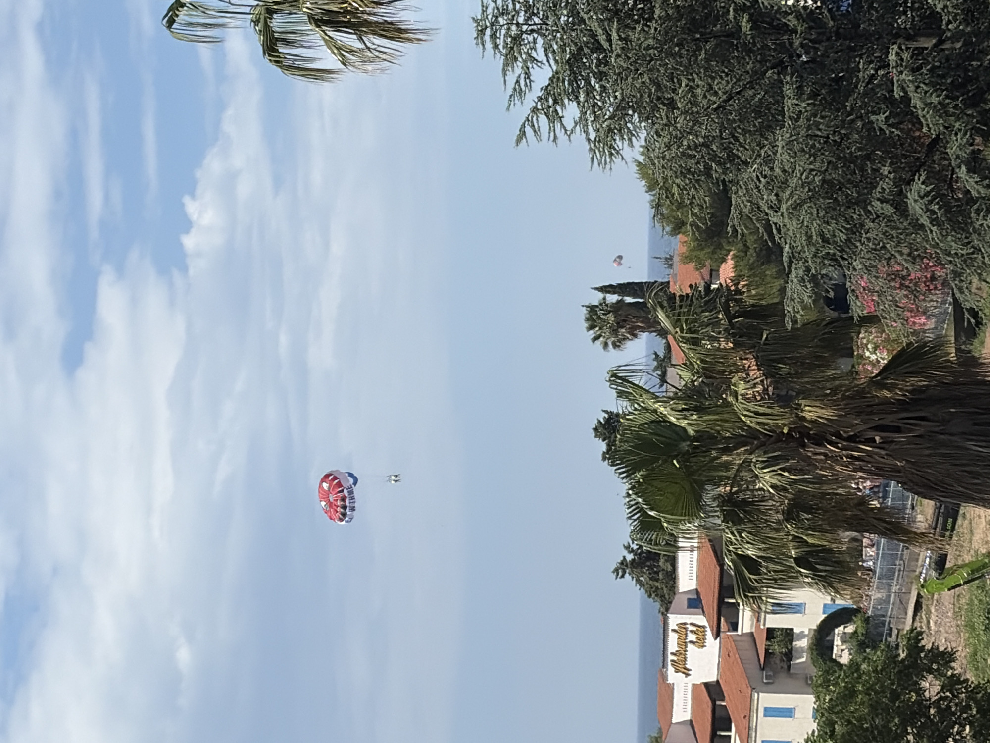 The image is a horizontally oriented, low-angle shot of a coastal scene. In the sky, a red and white parasail is visible with two people attached. Palm trees and other foliage flank the edges of the frame, partially obscuring the view of buildings with terracotta-tiled roofs. A building with "Aleksandar Hotel" written on it is visible in the bottom right, indicating the city of Budva, Montenegro. The sky is predominantly blue with scattered clouds.