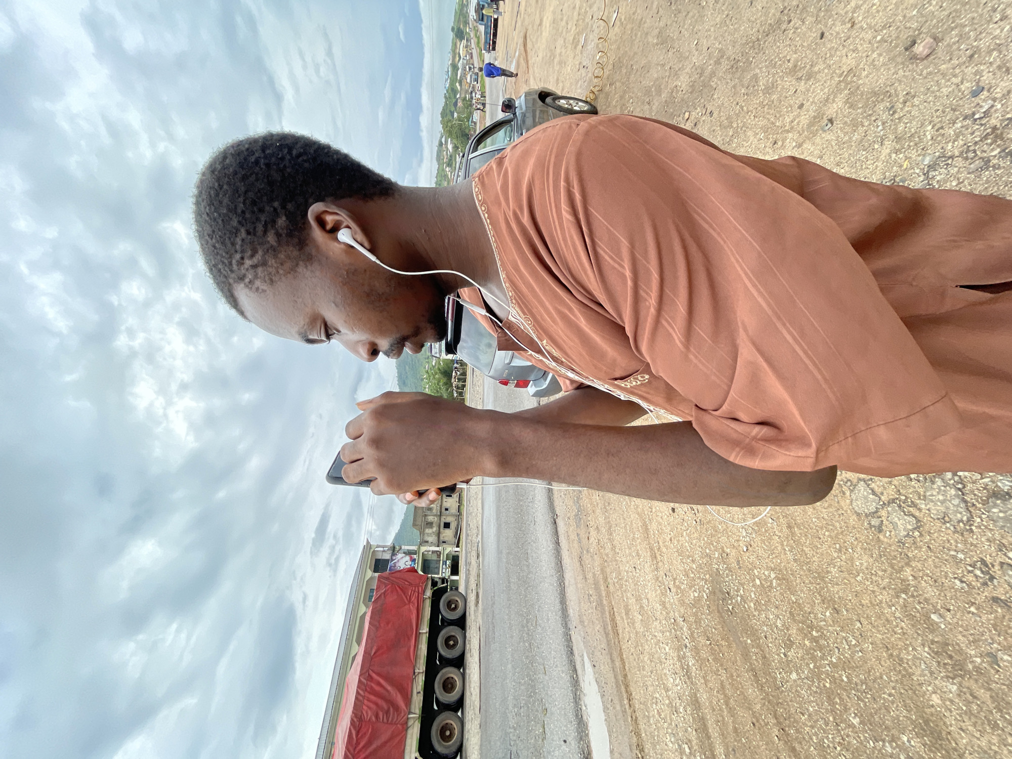 In Lokoja, Nigeria, a young man with short, dark hair is captured in a close-up, profile view, looking down at his phone and wearing white earbuds. He is dressed in a brown, patterned garment. The scene is outdoors, with a cloudy sky overhead. In the background, a large truck with a red tarp-covered load is visible, along with several cars and a distant, hazy view of buildings and vegetation. The ground is unpaved, composed of dirt and small stones, suggesting a rural or semi-developed area. The man's focus on his phone and the presence of earbuds imply he is engaged in communication or entertainment. The overall atmosphere is casual and everyday.