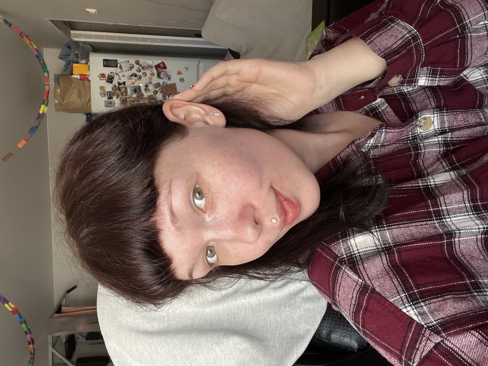 A woman with auburn hair and green eyes, wearing a burgundy and white plaid shirt, is lying on a white pillow. She has a stud piercing on the left side of her lower lip and a stud earring on her left ear. Her left hand is raised towards her ear. Behind her is a white refrigerator covered in magnets and photos, and a colorful toy arching across the wall. The location is not specified in the image, and there is no evidence to suggest it is Chaikovsky, Russia.