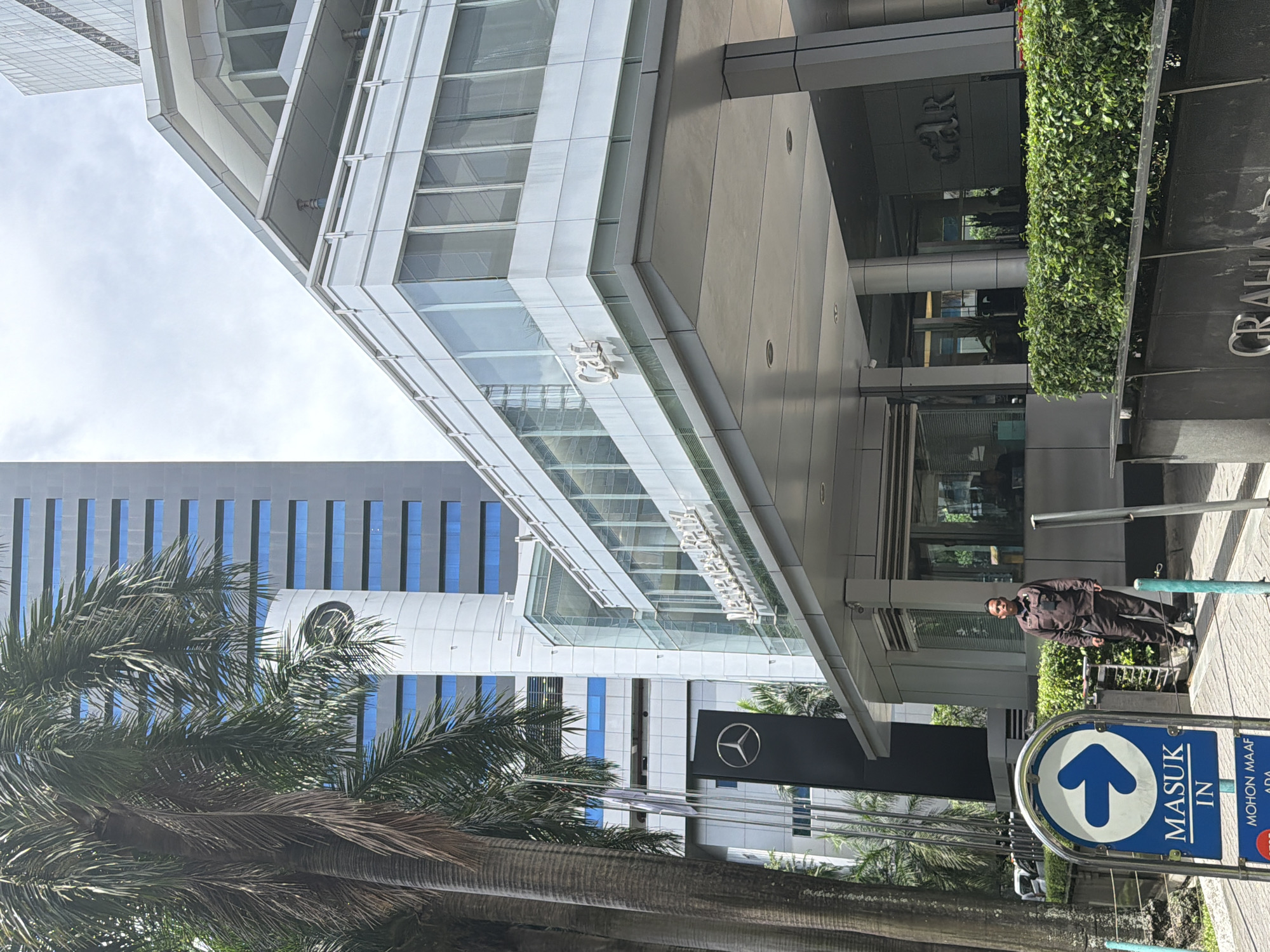 The image captures a street scene in South Jakarta, Indonesia, showcasing a modern building with glass and steel architectural elements. The building features signage indicating "GRAHA CIMB NIAGA," with a smaller "CIMB" logo visible on the glass facade. Adjacent to the building, there is a Mercedes-Benz dealership sign featuring the brand's iconic logo. The view is partially obstructed by palm trees in the foreground. A uniformed security guard is standing near the building's entrance, next to a blue and white sign that reads "MASUK IN," with an upward-pointing arrow. This suggests an entrance or access point. The weather appears bright and sunny, with shadows indicating the direction of sunlight.