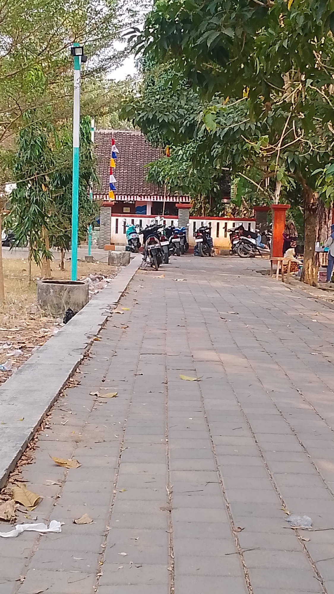 The image depicts an outdoor scene in Karanglangu, Indonesia, featuring a paved walkway lined with trees and a building with a tiled roof adorned with colorful flags. Several motorcycles are parked along the walkway in front of the building. The environment appears to be a public space, possibly a park or a community area, with fallen leaves and some scattered debris along the side of the walkway. The lighting suggests it is daytime.