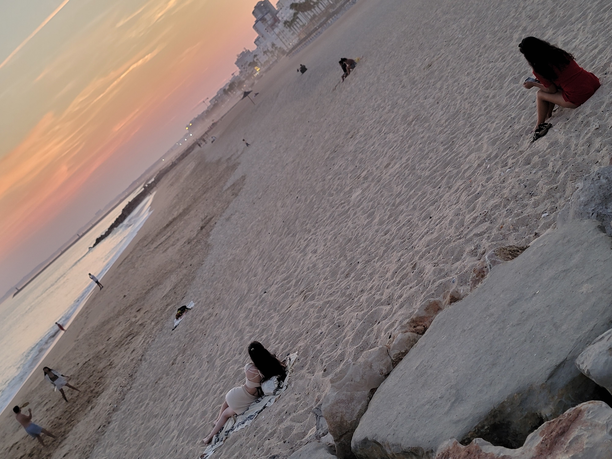At Quarteira Beach, Portugal, during sunset, several individuals are present on the sandy shore. In the foreground, a person in a red dress is seated on the sand, holding a phone. Further up the beach, a person is lying on a patterned blanket with their legs extended. In the mid-ground, a group of people are scattered across the sand, some walking, others sitting. The ocean is visible in the background, with gentle waves lapping the shore. The sky displays a gradient of warm colors, indicative of the setting sun, with wispy clouds streaking across. The horizon features the outline of buildings and structures, suggesting the presence of the town of Quarteira. Rocks are visible in the lower left portion of the frame, bordering the sandy area.