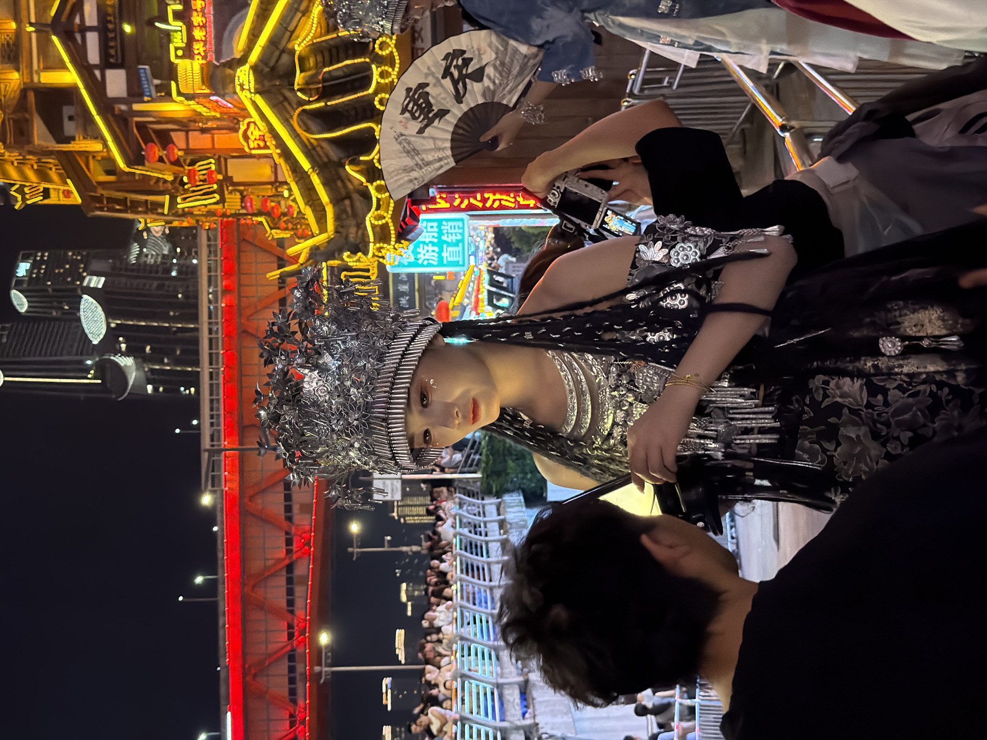 A woman dressed in traditional Chinese attire, including an elaborate silver headdress and a dark, floral-patterned dress, is the central subject of this nighttime photograph. She is looking towards the camera with a serene expression. Her attire is adorned with intricate silver embellishments and jewelry.
In the background, a bustling street scene unfolds, illuminated by vibrant neon signs and traditional Chinese architecture. The lights are bright yellow and red, suggesting a lively atmosphere. A white fan with black Chinese characters is visible, held by someone to the woman's left. A person is also seen holding a camera, possibly taking a photograph of the woman.
A red bridge structure is visible in the lower portion of the image, further contributing to the urban nightscape. The overall scene appears to be a street or a tourist area in Yuzhong District, China, during the evening or night. The presence of many onlookers suggests a performance, festival, or a popular tourist attraction. The mood conveyed is festive and visually rich due to the vibrant lights and detailed costumes.