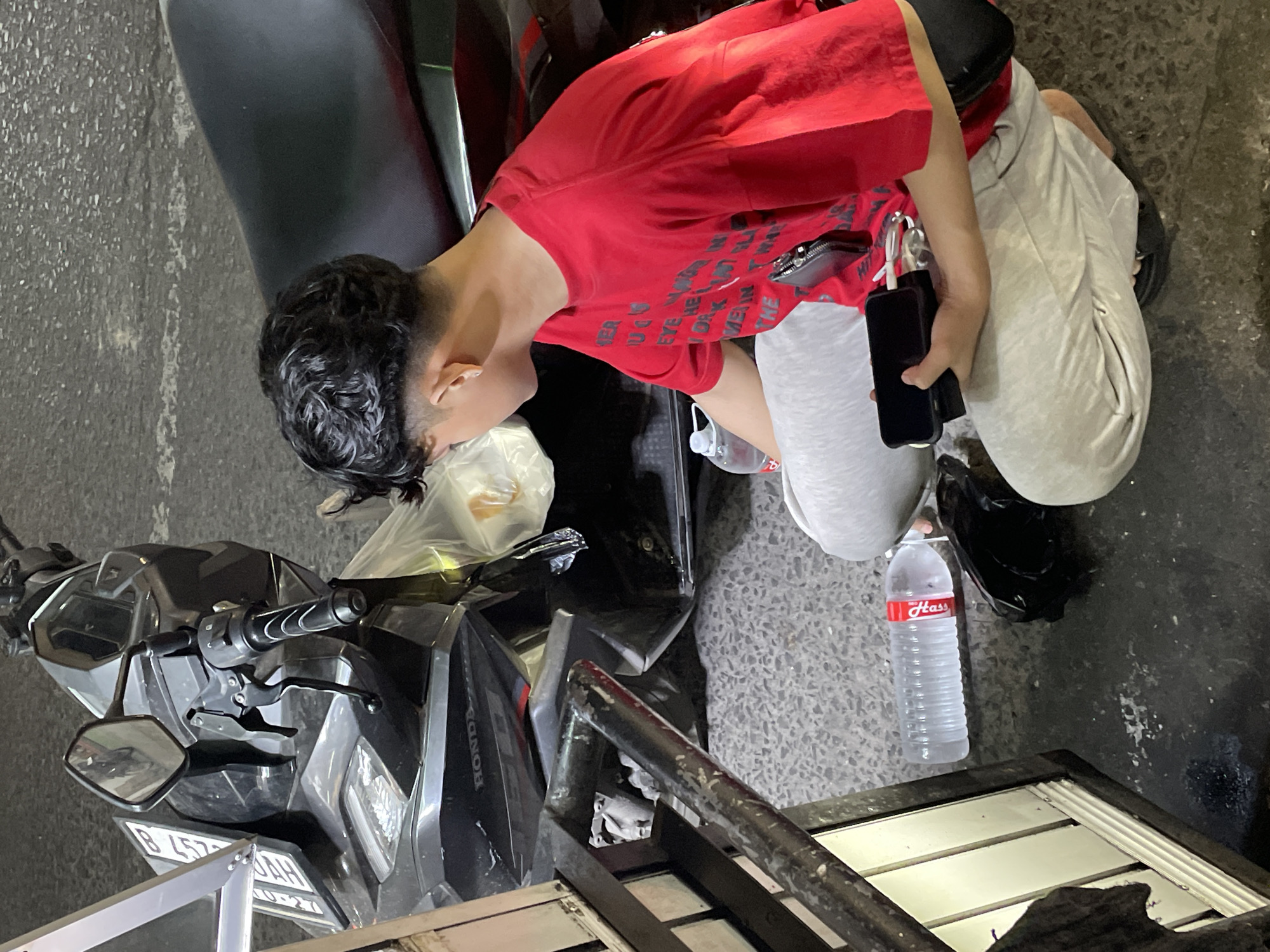 This media file shows a young boy with dark, curly hair wearing a red t-shirt and grey sweatpants. He is leaning over a black motorcycle. The boy is holding a black smartphone in his right hand and a portable charger is attached to it. He appears to be looking at something near the motorcycle. In his left hand, he is holding a clear plastic bag containing some food items. A clear plastic bottle of water with a red label is visible nearby. The scene appears to be outdoors, with a textured, greyish ground. The motorcycle is parked, and its headlights and handlebars are visible. The overall impression is that of a casual moment, possibly in a street or parking area. The lighting suggests it might be daytime, but the exact time is not discernible. The location is identified as North Jakarta, Indonesia. No specific emotional cues are readily apparent, though the boy's focus is on his interaction with the phone and the items he is holding. A license plate on the motorcycle is partially visible, showing the characters "B 4572 DAH". A logo on the motorcycle reads "HONDA". The water bottle has a label that appears to say "Hass".