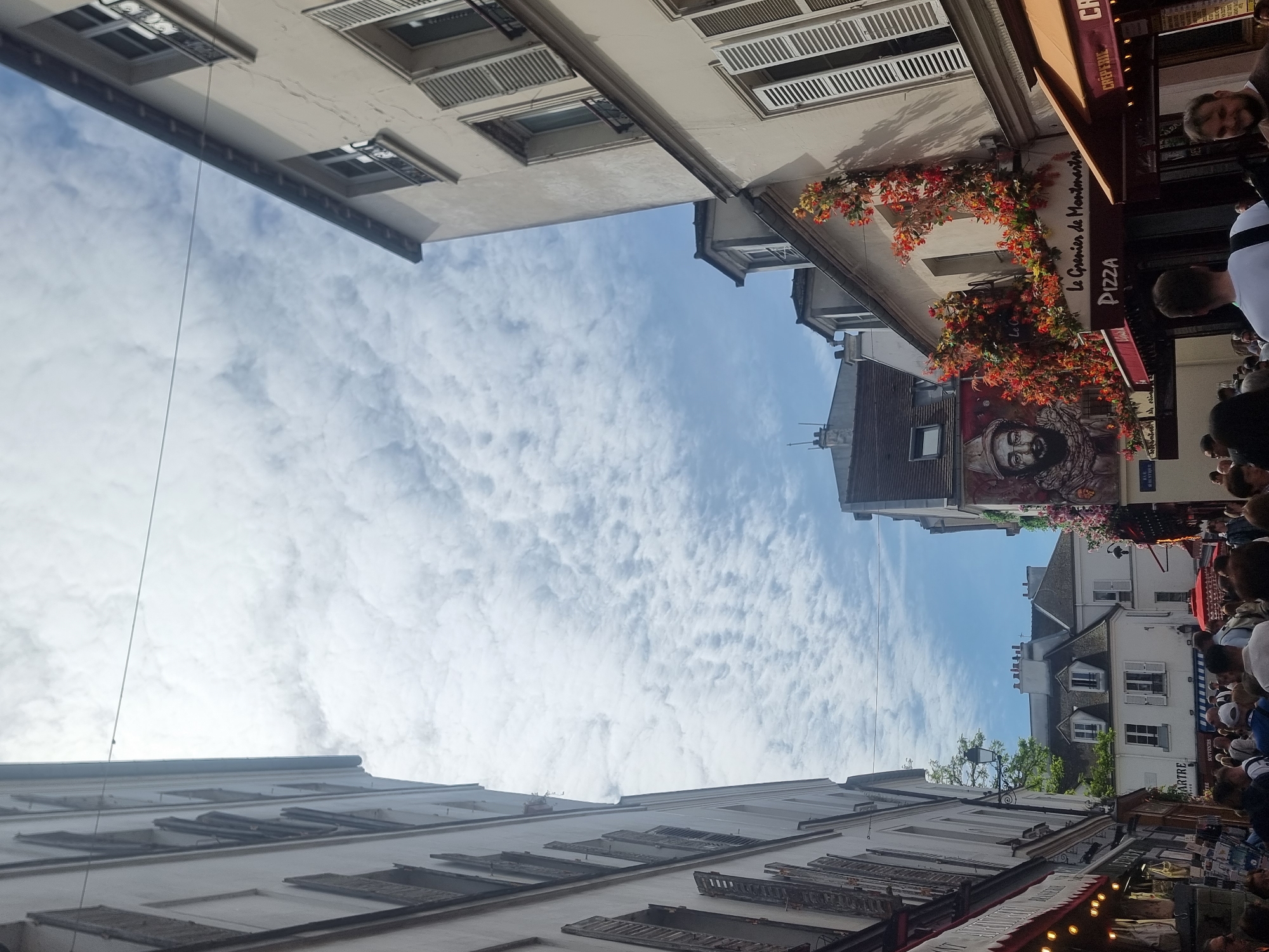 The image captures a vibrant street scene in what appears to be Montmartre, Paris, France, viewed from a low angle looking upwards. The original image is rotated 90 degrees clockwise.
**Subjects:**
The foreground and mid-ground are dominated by tall, light-colored buildings lining a narrow street. These buildings feature numerous windows, many equipped with traditional white louvered shutters, some open, some closed. One building on the right side of the street displays a large, colorful mural of a bearded figure, partially obscured by cascading artificial red and orange flowers and foliage. A crowd of people is visible at street level on the right, their heads and upper bodies seen beneath restaurant awnings, suggesting a bustling atmosphere. The sky, a bright blue with scattered white, wispy clouds, fills the upper central portion of the frame.
**Activity or Event:**
People are gathered on the street, likely walking, dining outdoors, or socializing. The presence of restaurant signs and awnings indicates commercial activity, characteristic of a lively pedestrian area.
**Notable Details:**
The scene takes place during bright daylight, suggesting a clear day in the mid-morning or afternoon. The architecture, including the multi-story buildings, ornate windows, and shutters, is typical of Parisian streets. A thin wire or cable runs vertically through the middle of the sky. The overall impression is one of a busy, pleasant urban environment.
**Visible Text:**
Several signs are visible on the buildings, including "Le Grenier de Montmartre," "PIZZA," and "CRÊPERIE." Another sign partially reads "MARTRE," further confirming the Montmartre location, and "BOUTIQUE" is also faintly visible.
